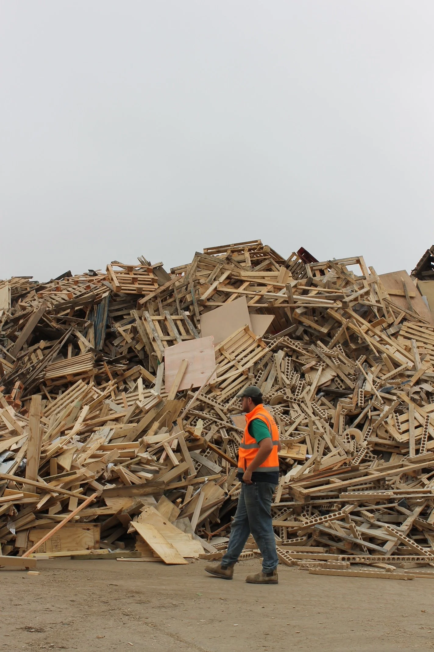 A person in a safety vest and boots walking past a large pile of wooden pallets and debris, with a cloudy sky above.
