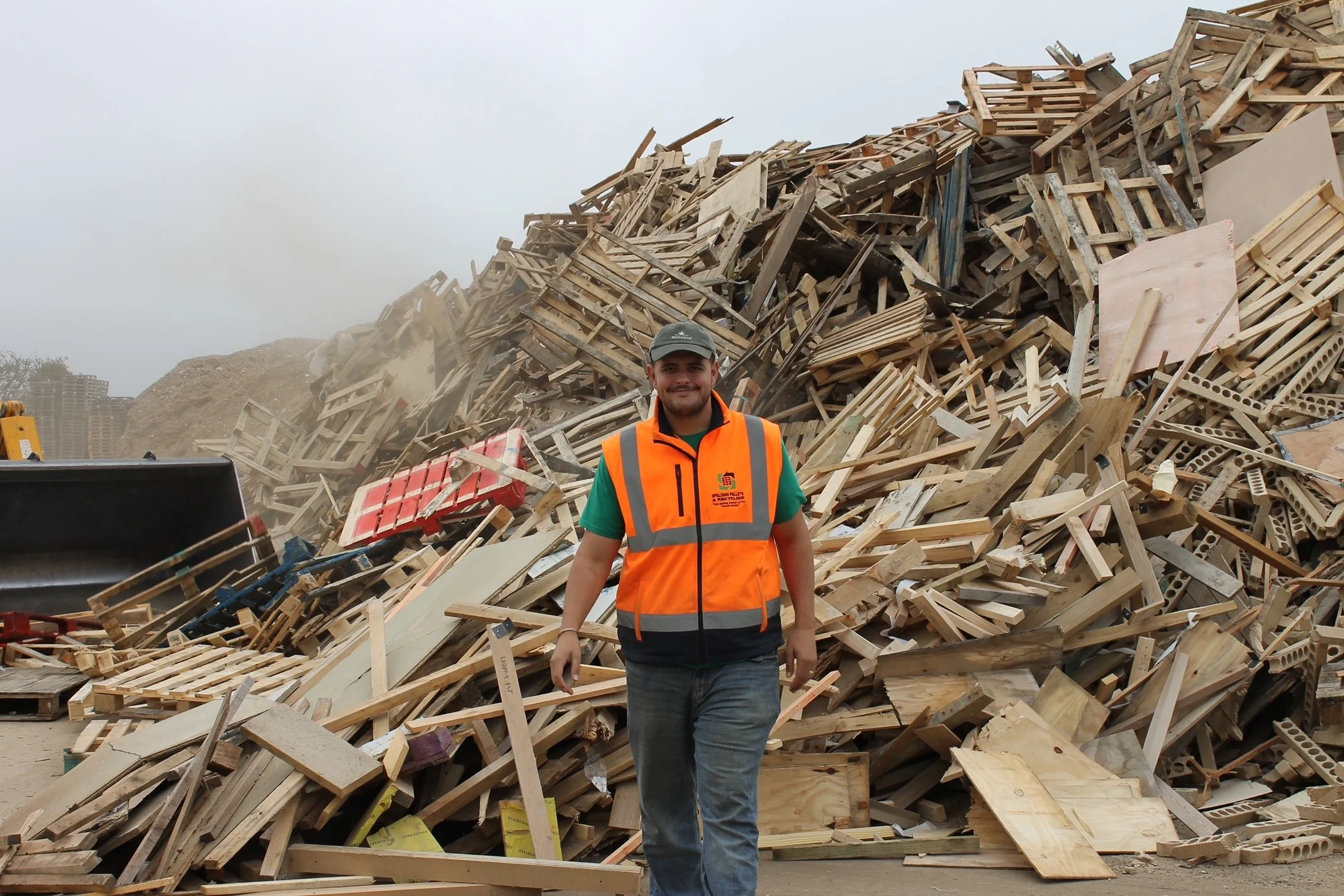 A man wearing an orange safety vest and cap stands in front of a large pile of wooden debris, including various scraps of lumber and building materials, at a construction or demolition site.