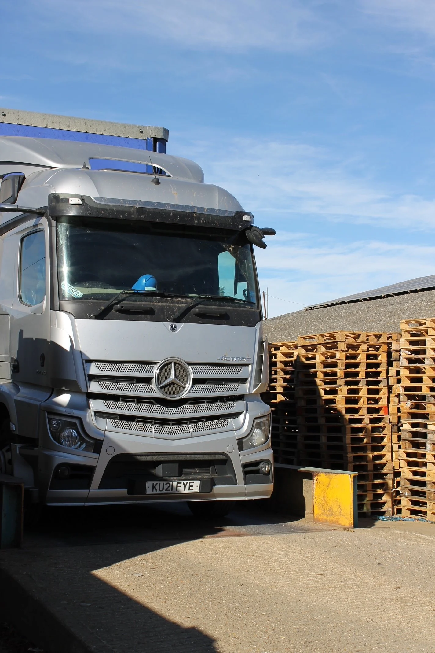 A silver Mercedes-Benz Actros truck parked next to stacked wooden pallets and a yellow barrier, with a blue sky and some clouds in the background.