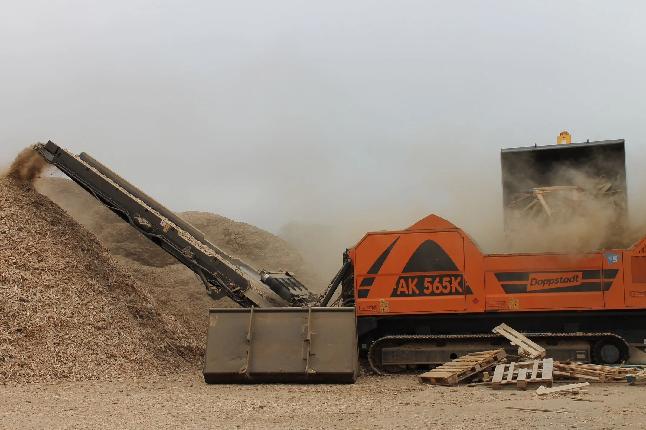 A conveyor machine transferring wood chips into a large container at a construction site with dust and wood pallets nearby.