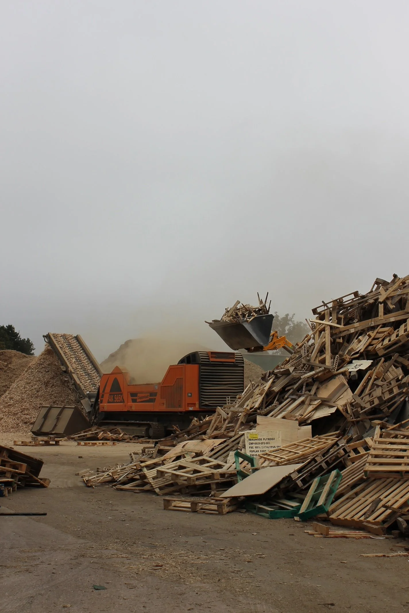 Large industrial wood shredder machine processing a pile of wood scraps at a construction or demolition site. The sky is overcast and there are scattered wood pieces on the ground.