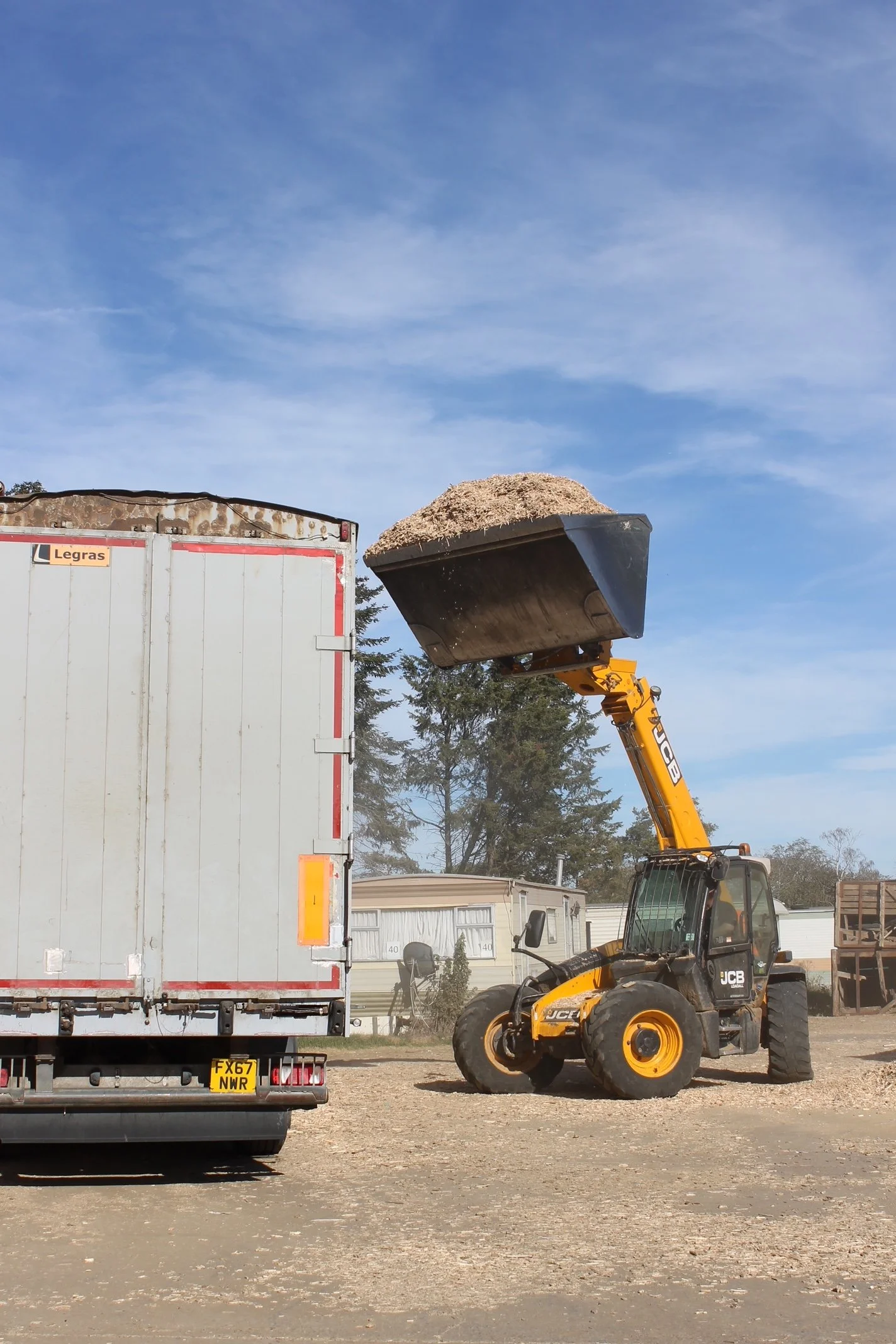 A construction site with a yellow JCB telescopic handler lifting a large load of sand into a truck. The scene includes several mobile homes in the background and a partly cloudy sky.