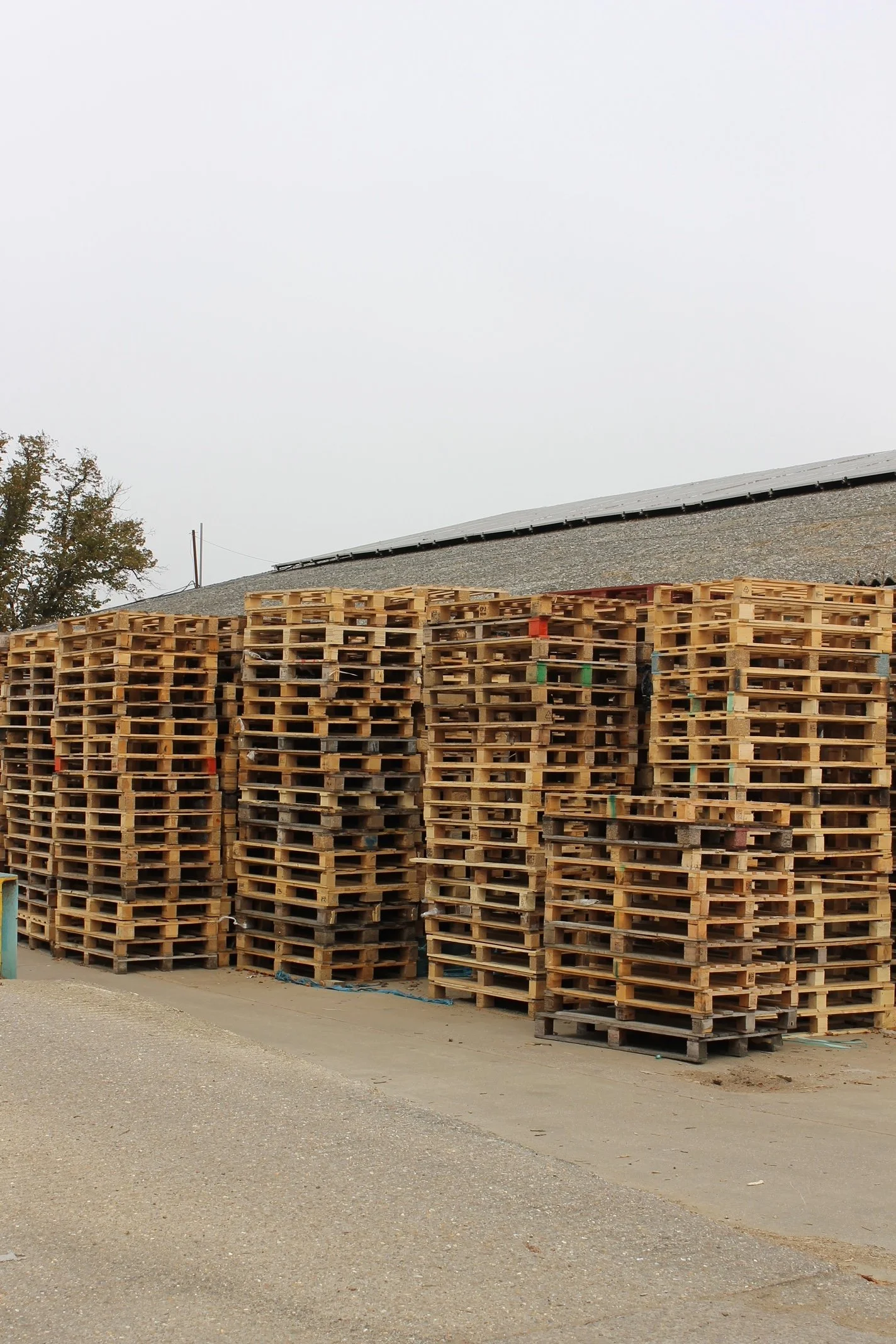 Stacked wooden pallets outdoors in front of a building with a sloped roof, overcast sky