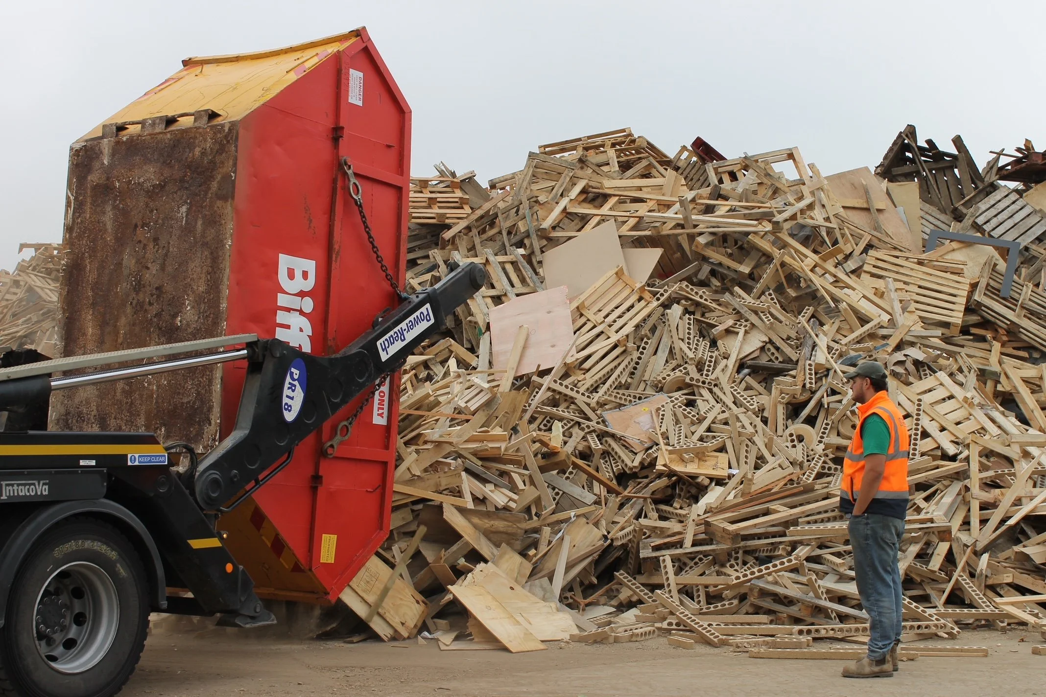 A large red dumpster truck dumping a huge pile of wooden pallets and scrap wood. A man wearing a safety vest and helmet stands nearby, observing the scene.