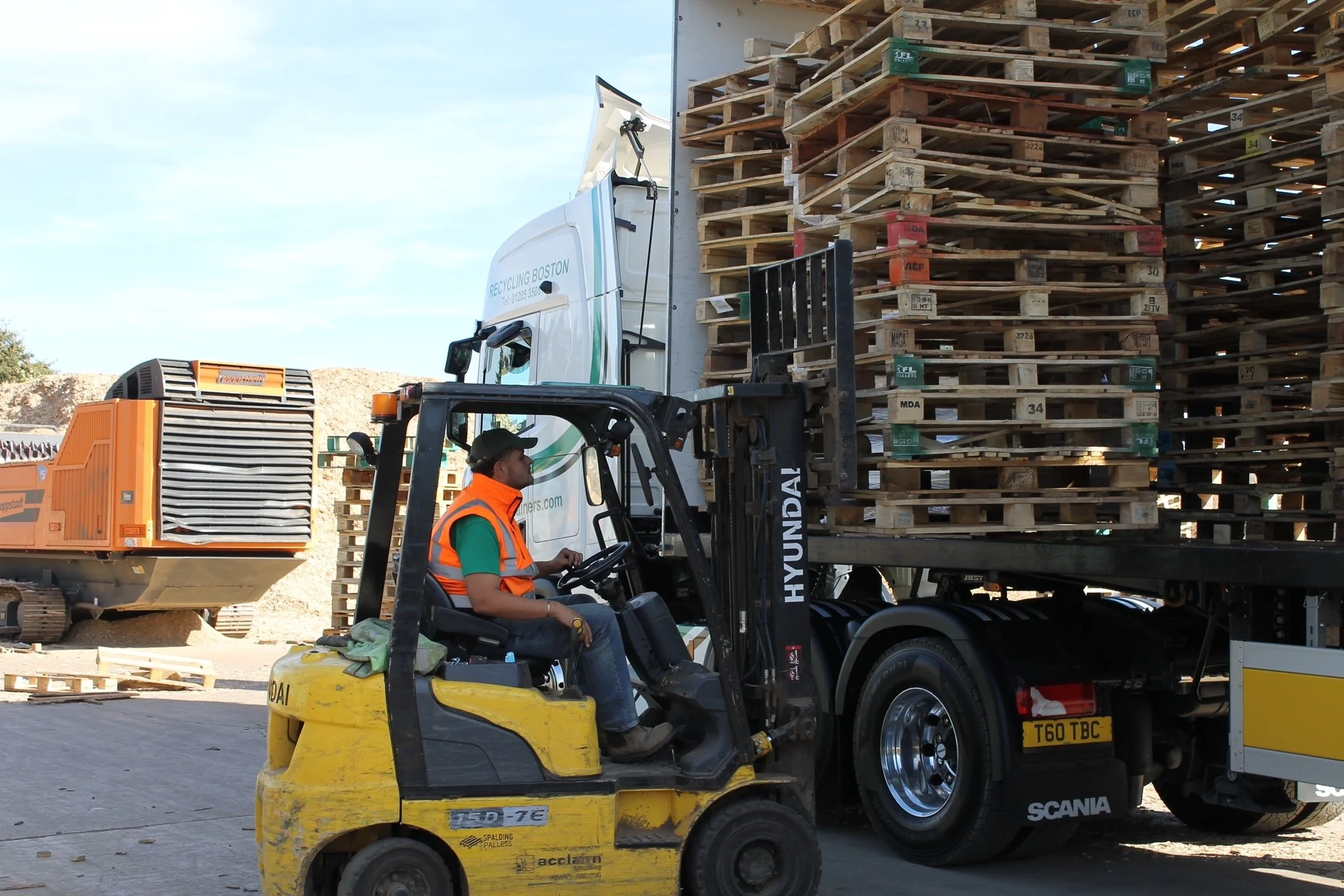 A forklift operator moving a pallet at a construction or recycling site with stacked pallets and construction equipment in the background.