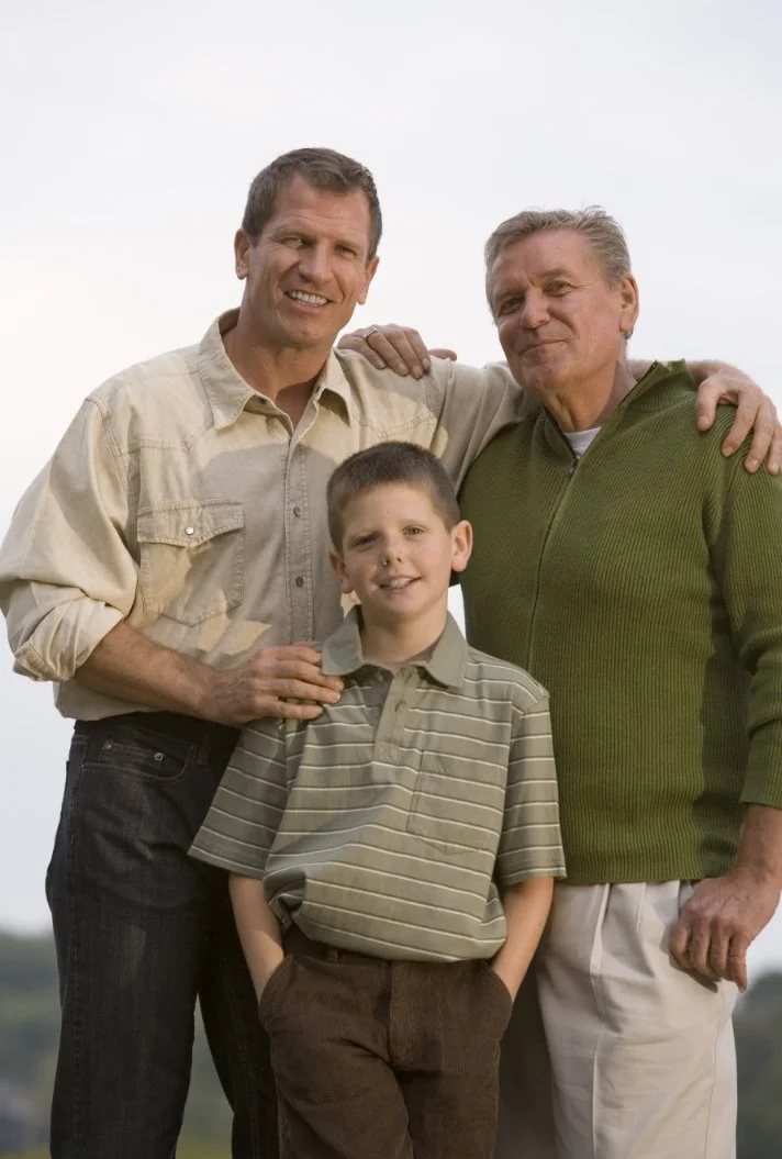 Three generations of men from the same family standing together outdoors, smiling, with one adult man placing his hand on the young boy's shoulder.