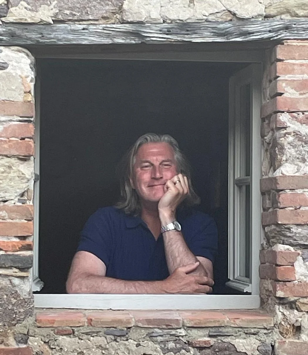 A man with long gray hair smiling while resting his chin on his hand, looking out of a window in a brick wall house.