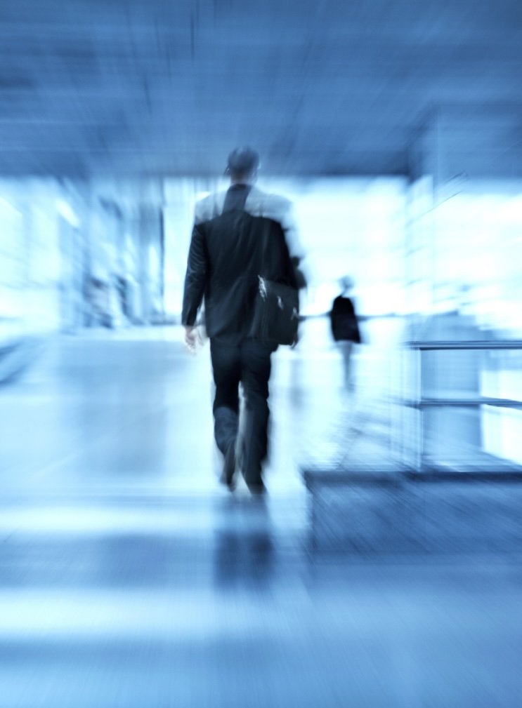 Man in black suit walking away in a modern, glass-walled building with another person in the background