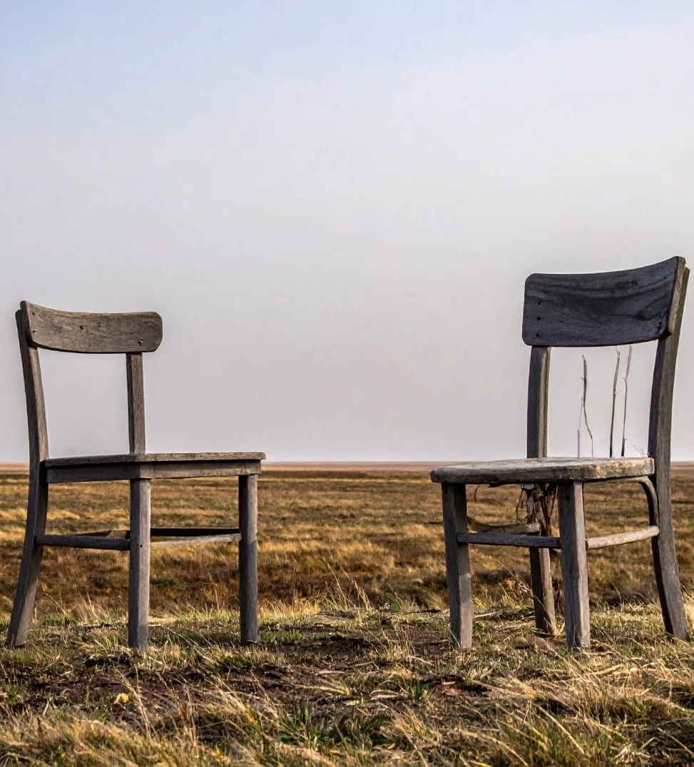 Two weathered wooden chairs facing each other in an open field with dry grass, under an overcast sky.