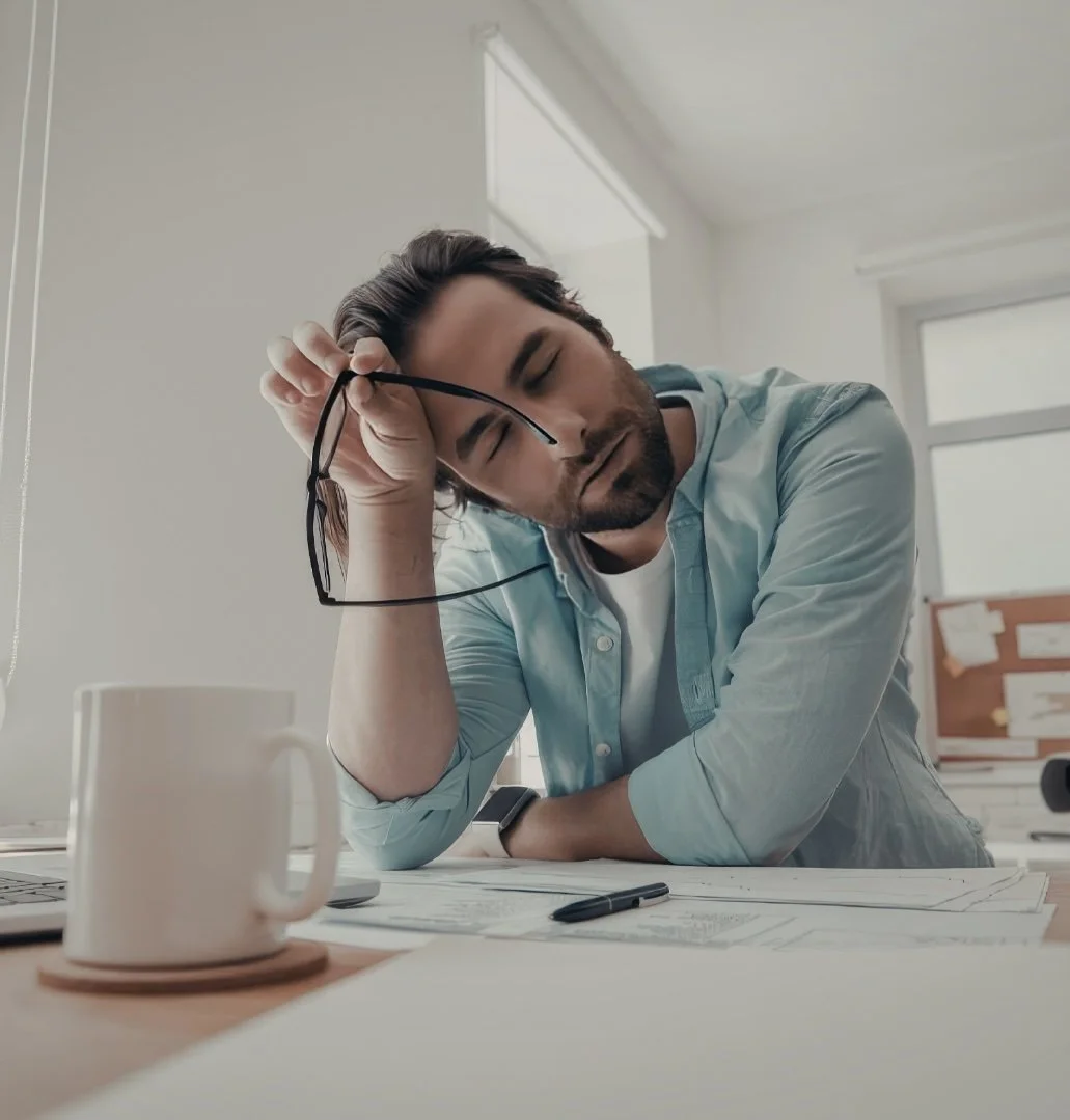 A man with dark hair and a beard, sitting at a desk with papers and a pen, holding a pair of glasses and resting his head on his arm, appearing tired or stressed in an office setting.