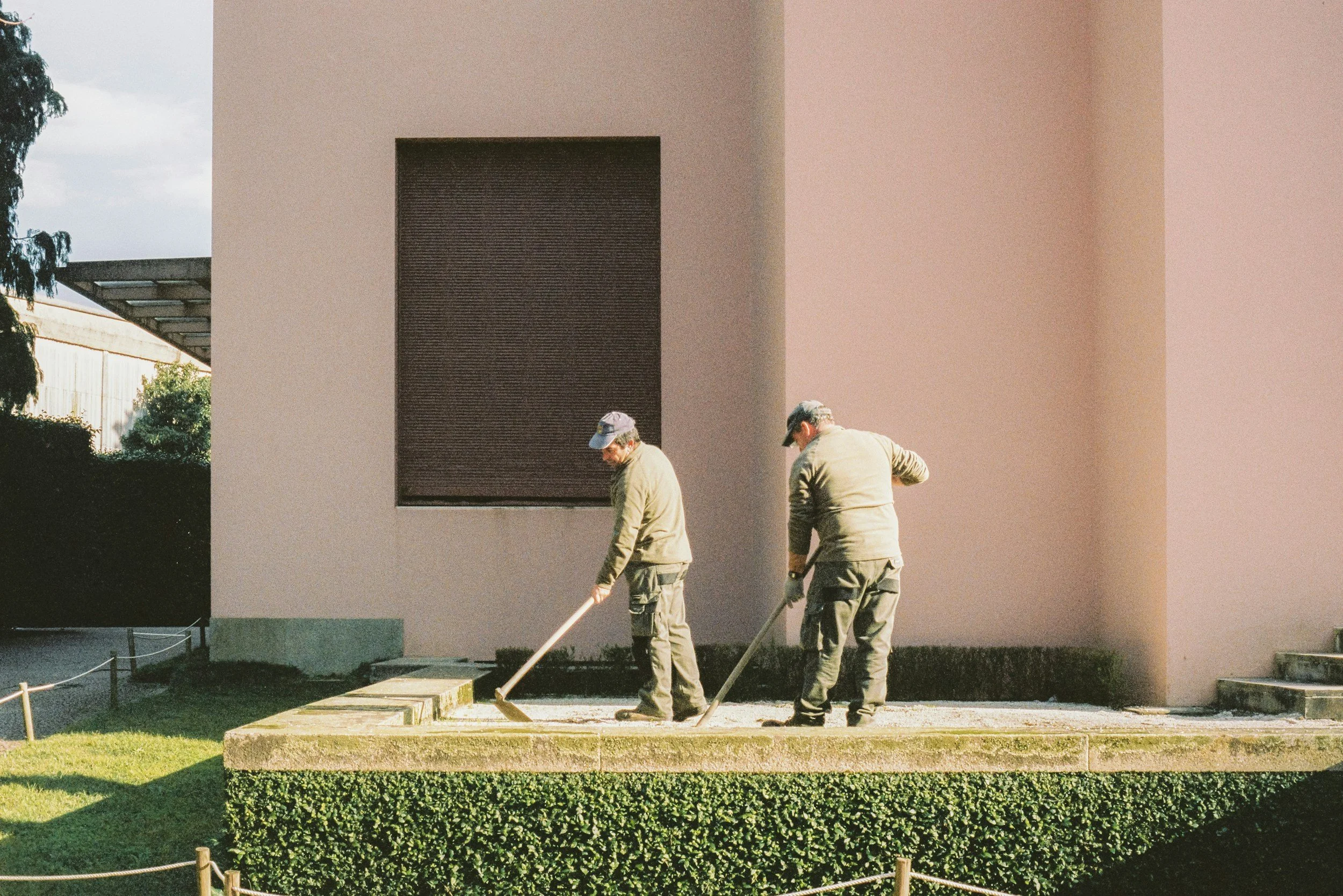 Two workers in beige uniforms and caps are working outdoors on a construction or landscaping project near a large modern building with a rectangular window. They are using tools to work on a rectangular stone or concrete surface.