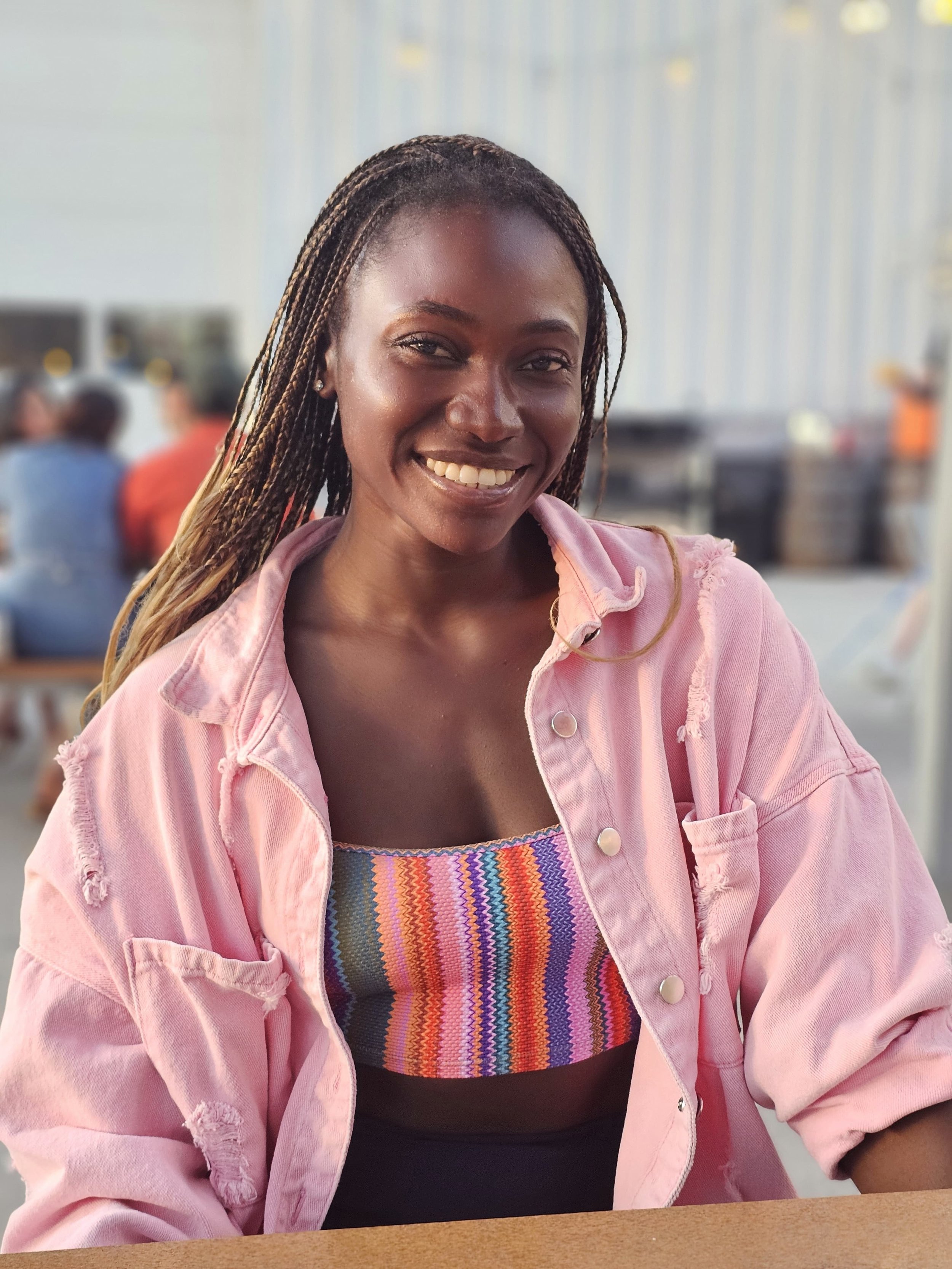 A woman with braids smiling, wearing a pink jacket over a colorful striped top, sitting at a table indoors.