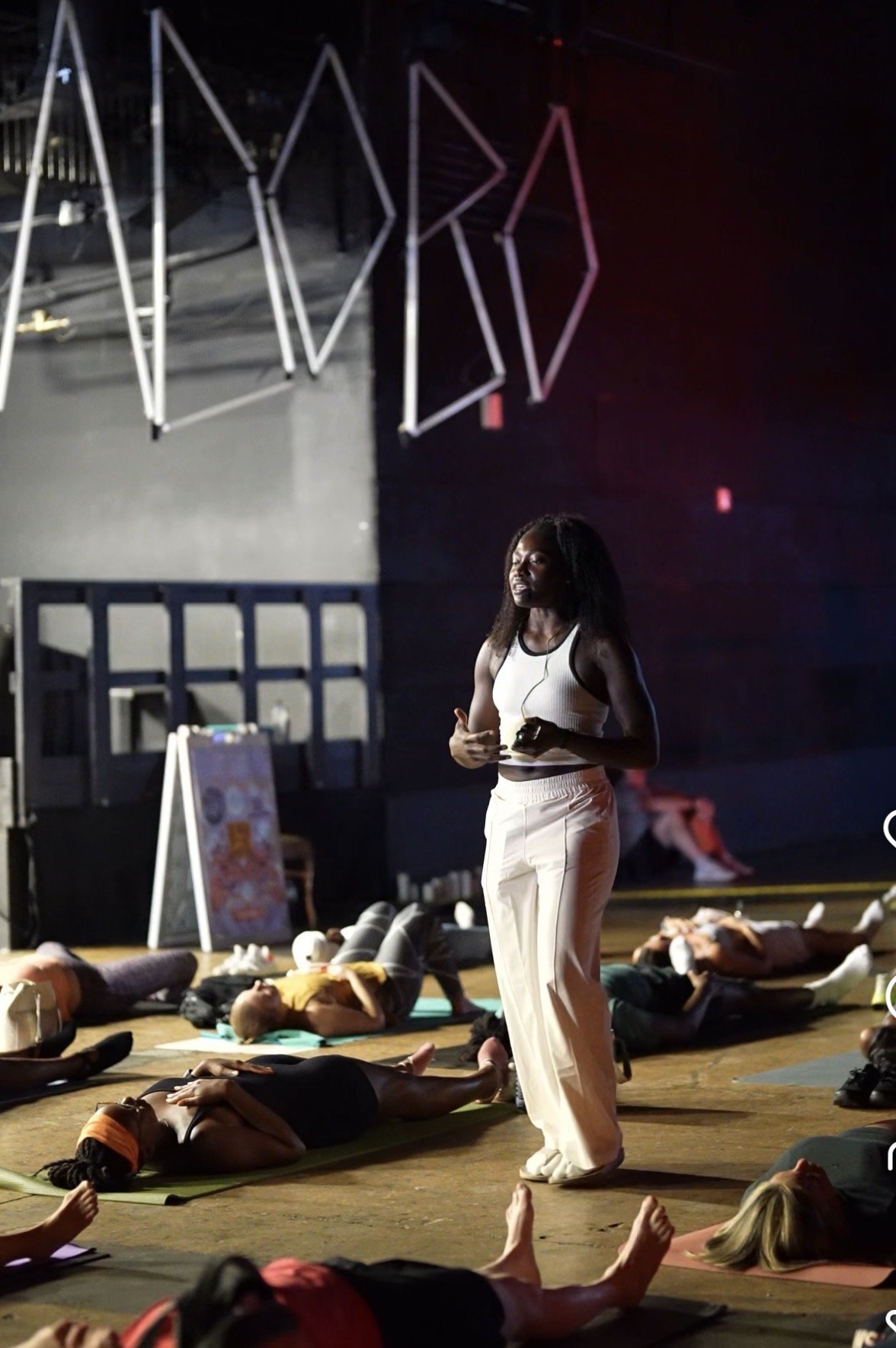 A woman leading a yoga class in a dark studio with black walls, illuminated by red and purple stage lighting, surrounded by people lying on yoga mats practicing yoga.