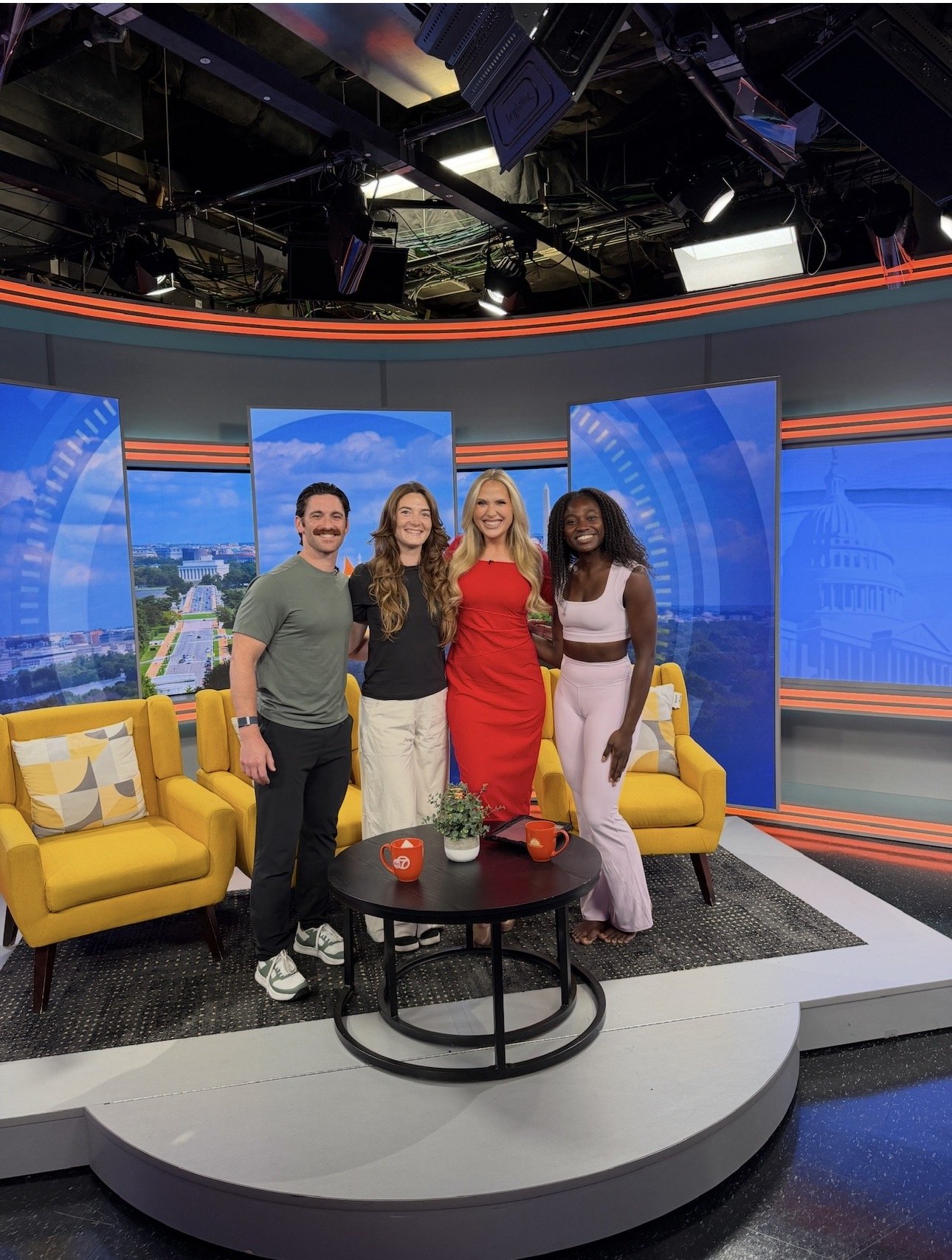 Four people standing together in a TV studio with a large screen behind them displaying a view of Washington, D.C. The group includes two women and two men, smiling and posing for the photo. The studio has yellow couches, a coffee table with two orange mugs and a small plant, and professional lighting and camera equipment overhead.