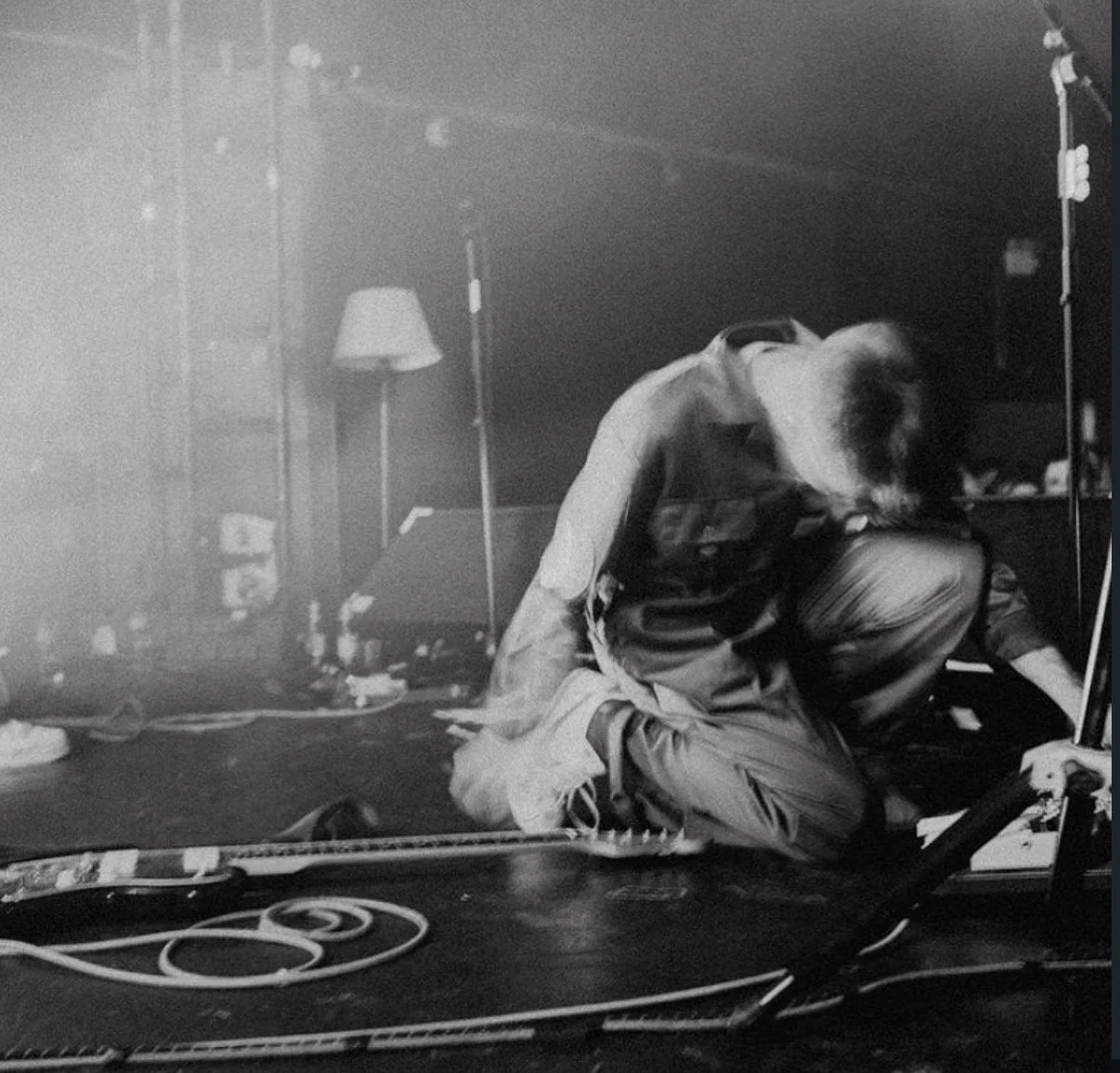 A person kneeling on the floor, head bowing down, in a cluttered room with a lamp, scattered papers and cords, and a microphone stand.