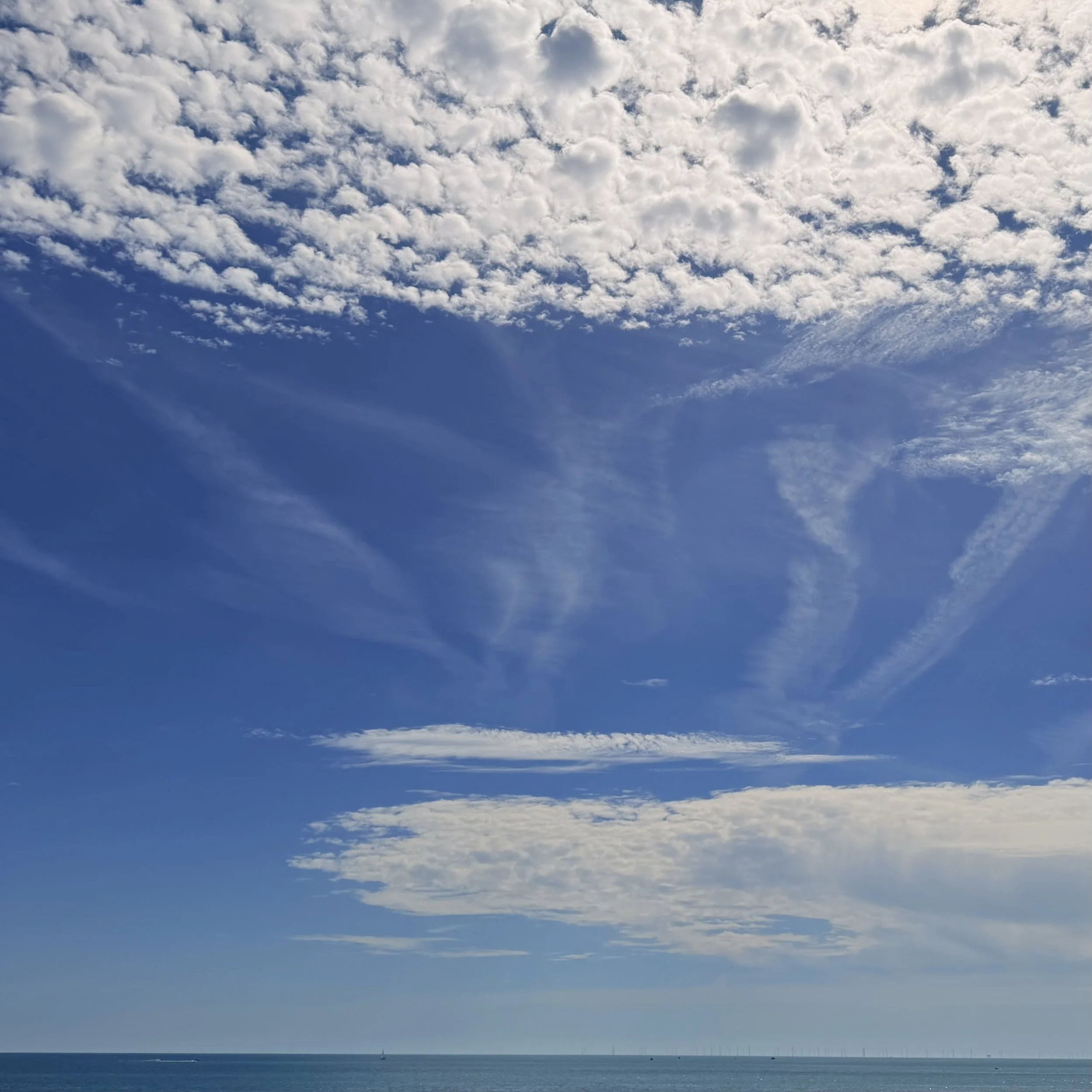 A large blue sky with scattered white clouds above a calm ocean, with several boats and wind turbines on the horizon.