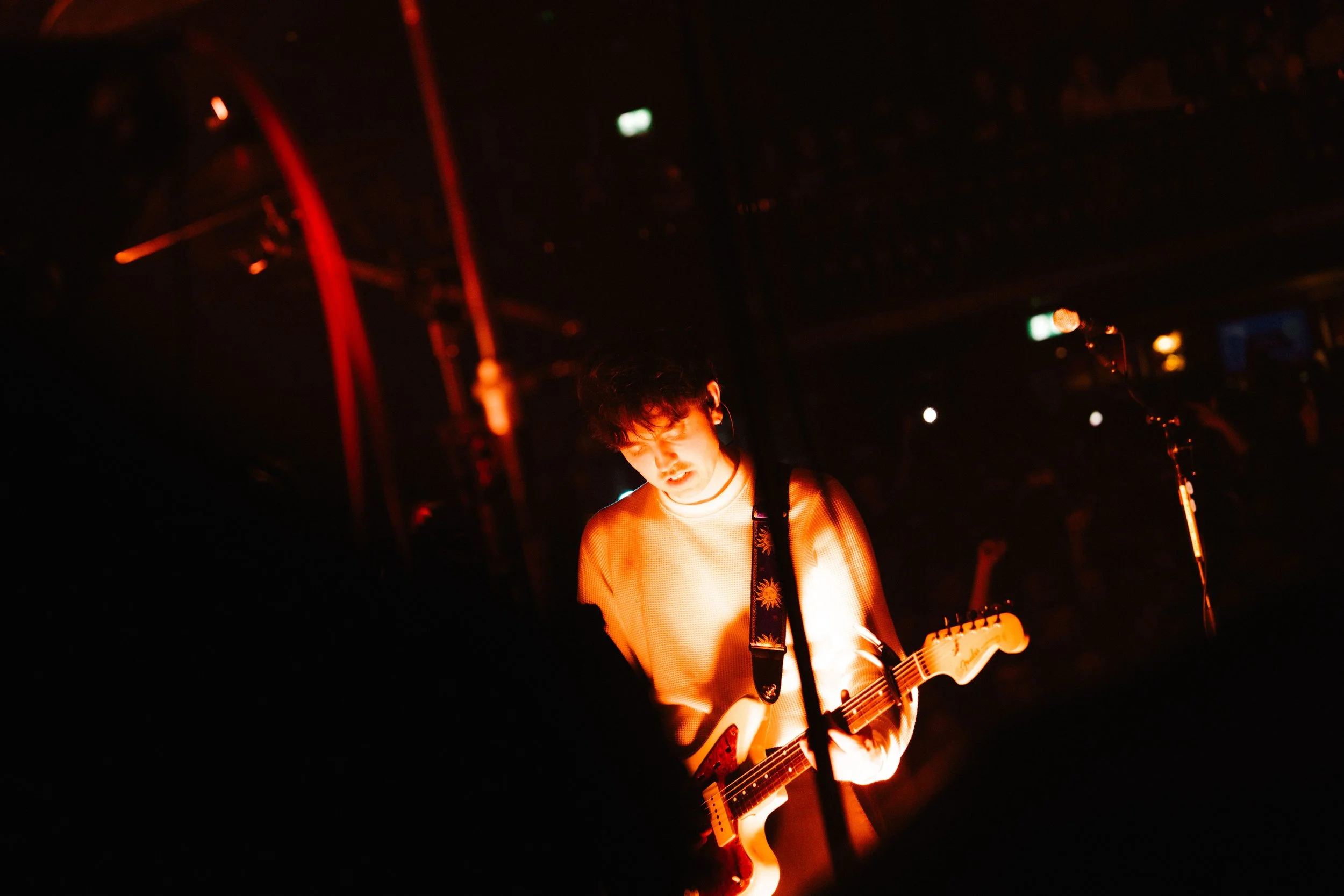 A musician playing an electric guitar on stage in a dimly lit environment with red and orange lighting, with a focused expression on his face.