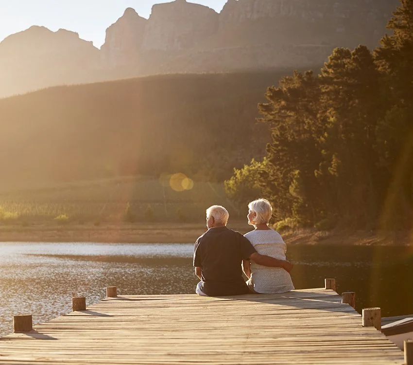 Deux personnes âgées assises sur un pont en bois au bord d'un lac, regardant le coucher de soleil dans un paysage naturel avec des montagnes et des arbres.