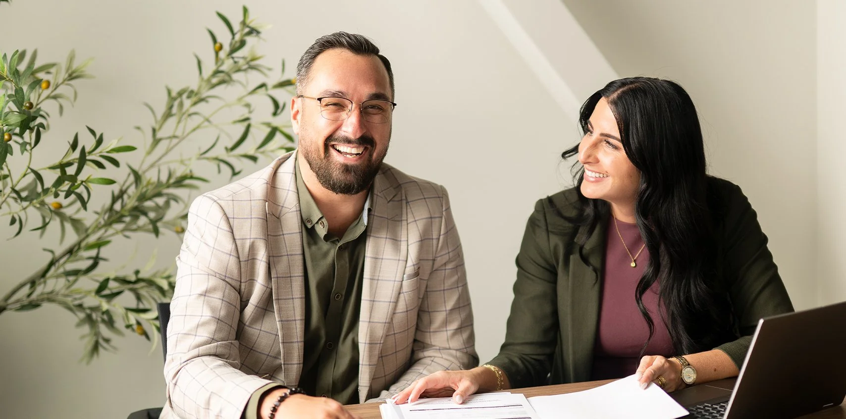 Deux personnes d'âge adulte souriantes en réunion, un homme avec une barbe, lunettes et veste à carreaux, une femme avec des cheveux noirs longs et une chemise violette, assis à une table avec documents et un ordinateur portable, dans un bureau éclairé