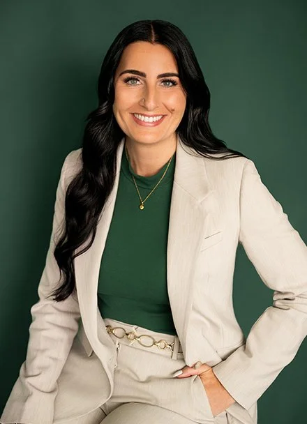 Femme souriante avec cheveux longs noirs, portant un blazer blanc et un haut vert, pose devant un fond vert.