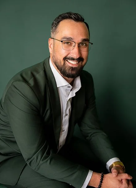 Portrait d'un homme souriant avec lunettes, portant un costume noir et une chemise blanche, assis contre un fond vert.