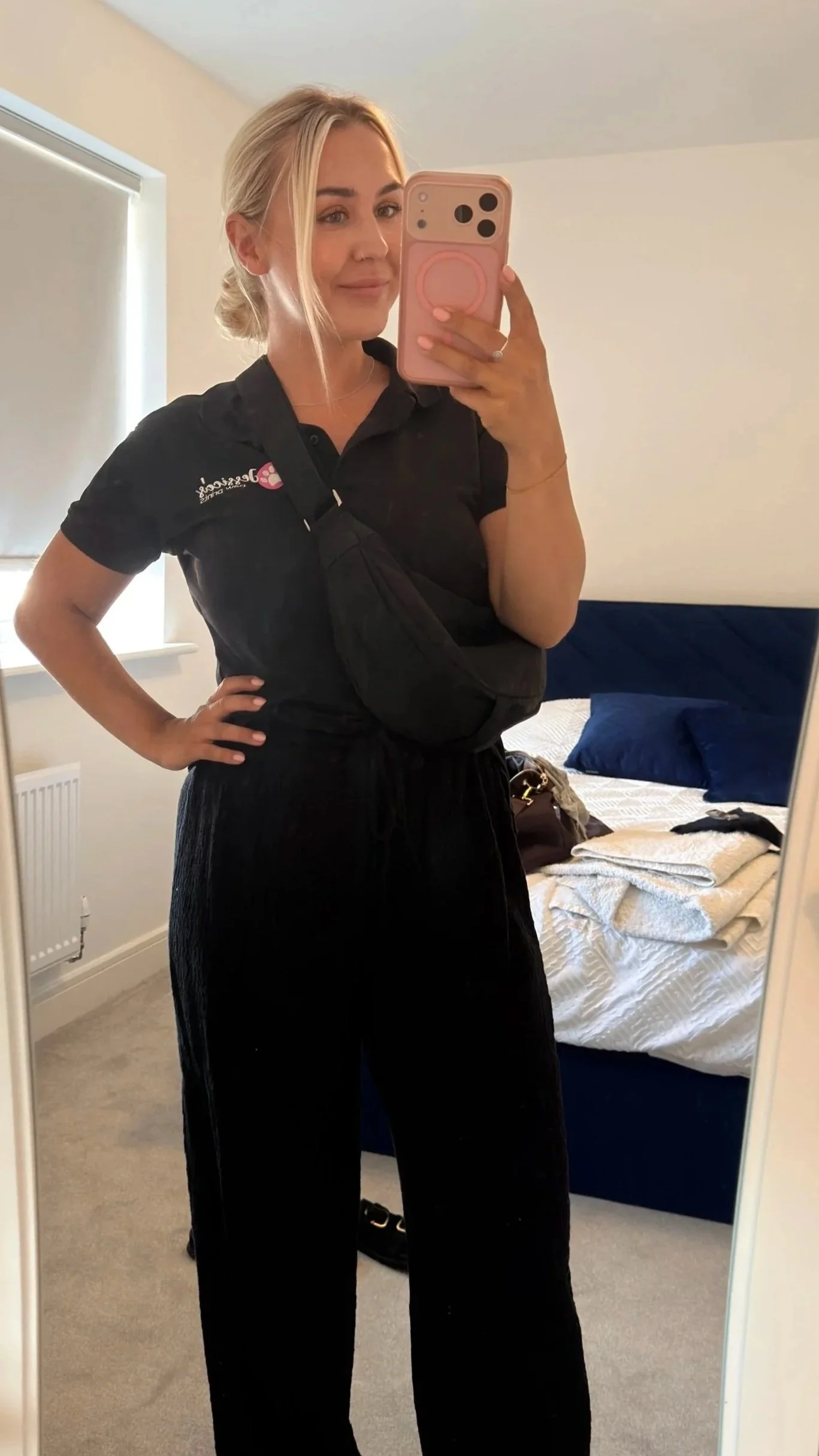 A woman with blonde hair taking a mirror selfie in a bedroom. She's wearing a black polo shirt and black pants, with a baby carrier strapped to her chest. The bed behind her has a dark blue headboard and is unmade, with a blue pillow, a brown bag, and some clothes on it.