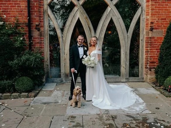 Bride and groom standing outside in front of a church entrance with large arched windows, holding a bouquet of white flowers, with a small dog on a leash.