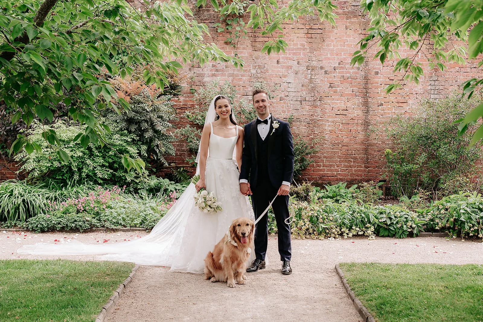 A smiling bride and groom in wedding attire holding hands and standing in a garden with a dog, flowers, and a brick wall background.