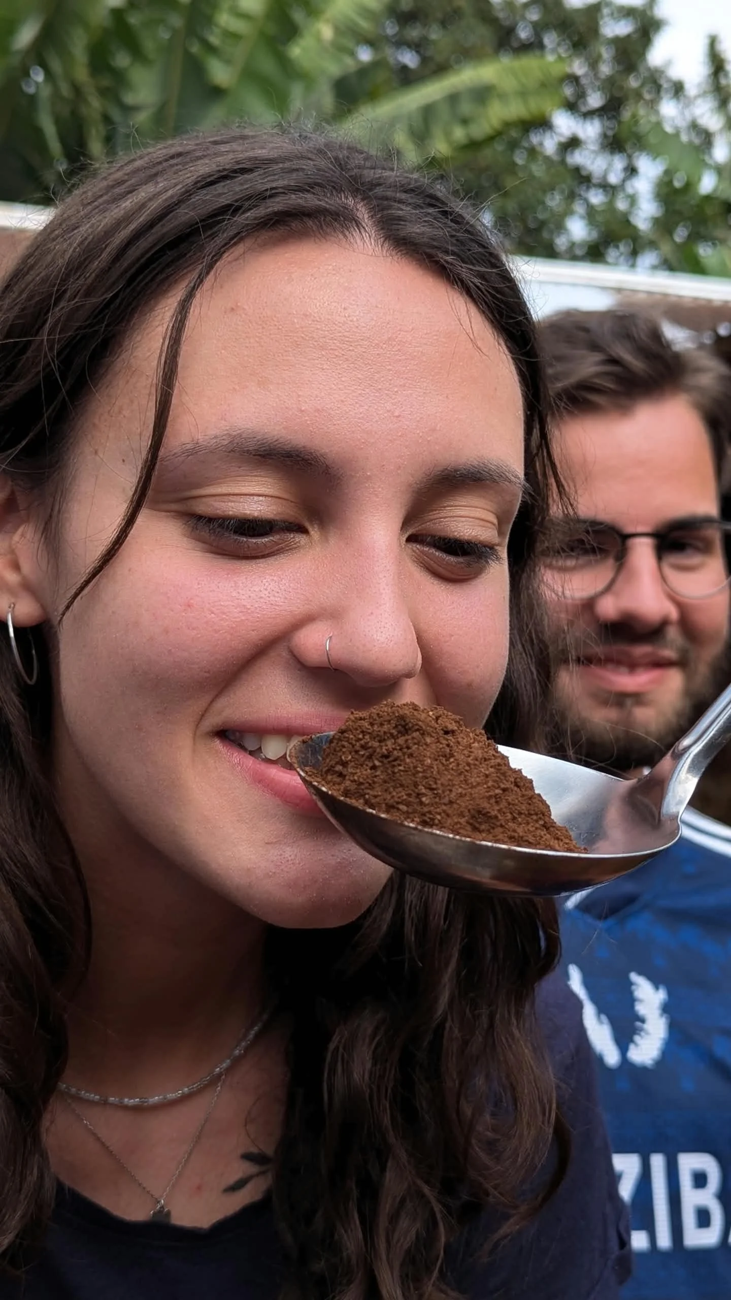 A woman with long dark hair and a nose ring holding a spoonful of ground coffee close to her mouth, smiling slightly. A man with glasses and brown hair is in the background with a slight smile, outdoors with green foliage.