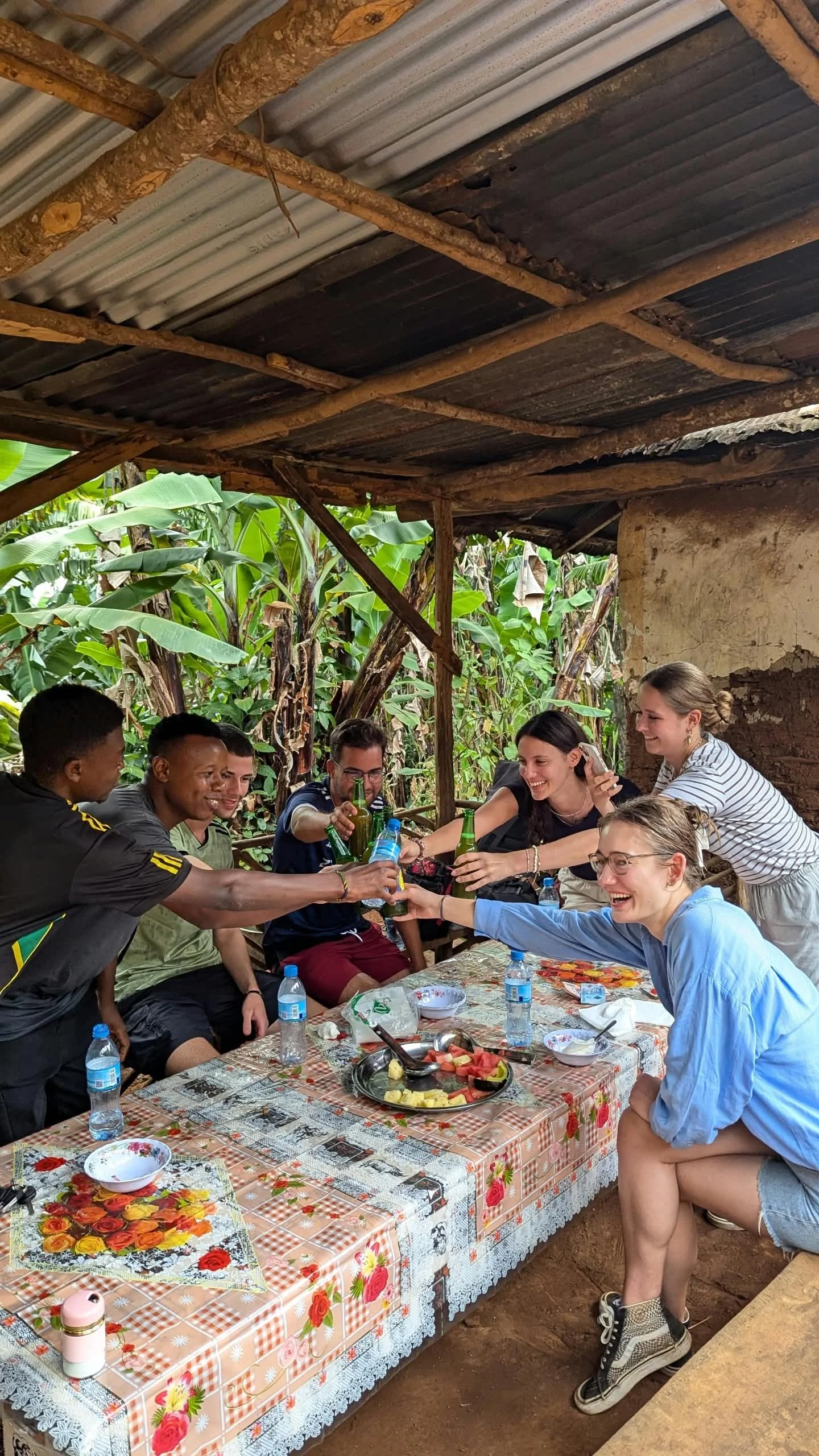 Group of people sitting at a table outdoors, raising bottles of drinks for a toast, with a covered rustic structure and lush green plants in the background.