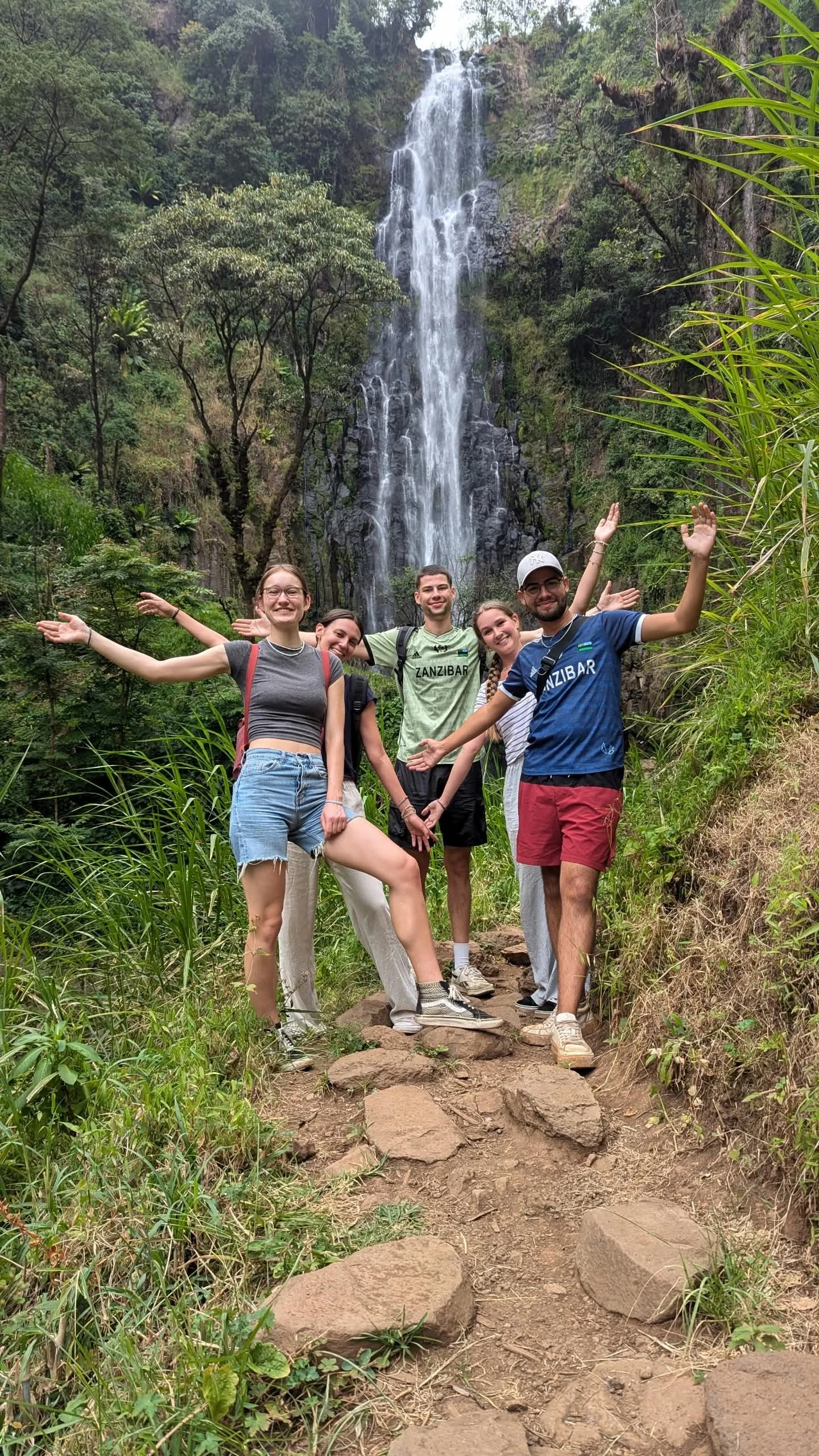 Group of five happy young adults posing with arms raised in front of a lush green landscape with a waterfall cascading down a rocky cliff, surrounded by trees and plants.