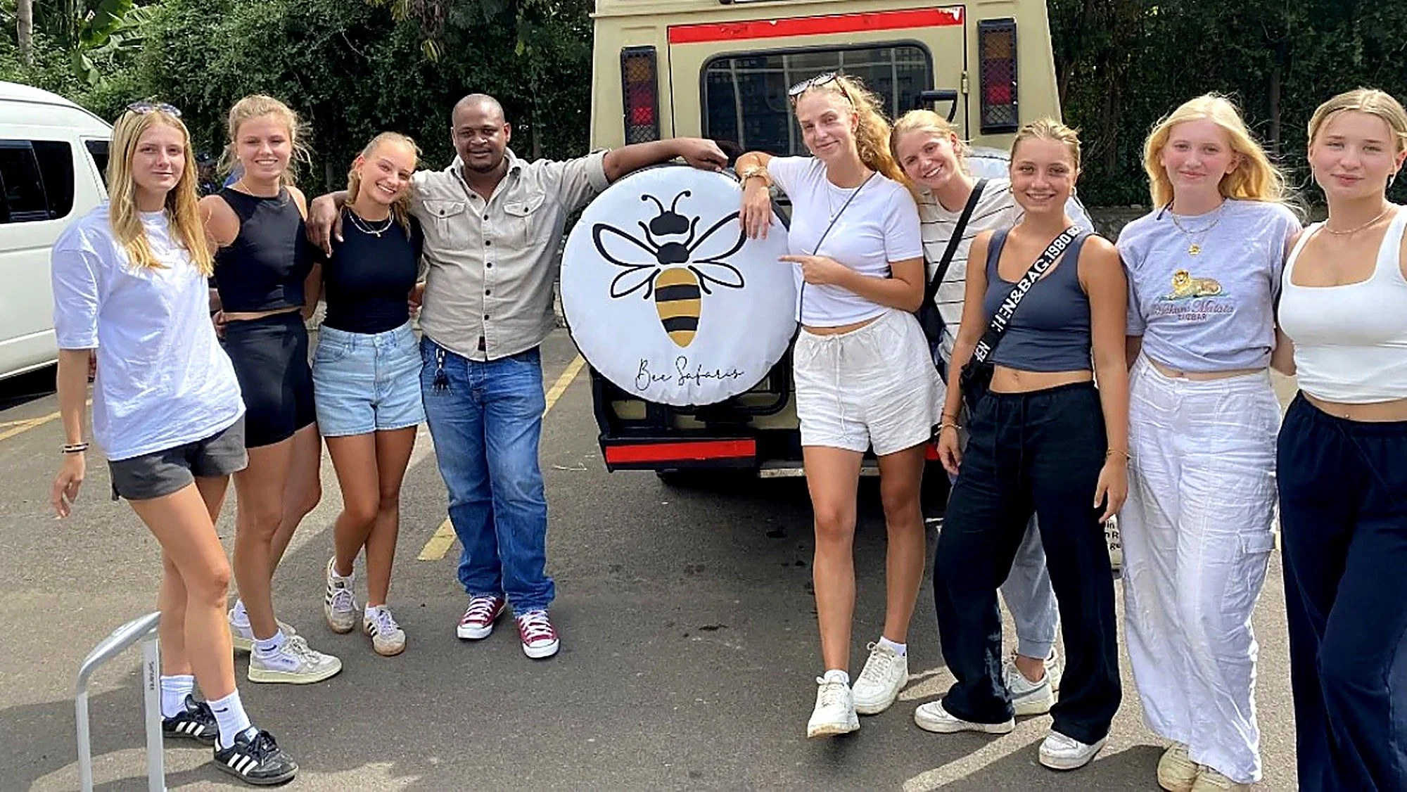 A group of nine young women and one man standing outdoors next to a bee-themed sign with a bee graphic and the text 'Bee Safely.' They are smiling and casually dressed, with some wearing sunglasses and sneakers. Behind them are parked cars, a large vehicle, and greenery.