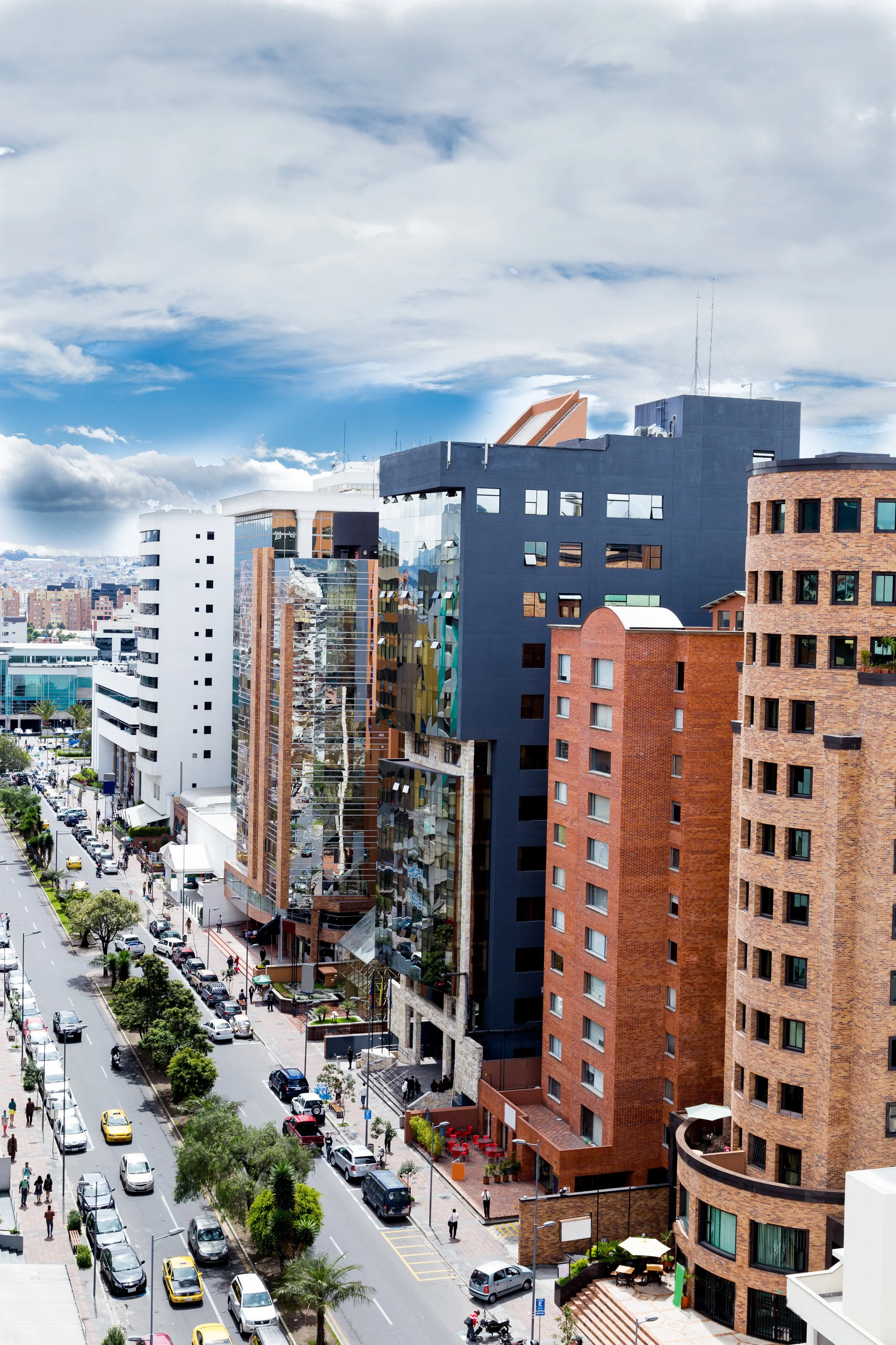 A busy city street with cars and pedestrians, surrounded by modern multi-story buildings including a black and red brick building, with a partly cloudy sky overhead.