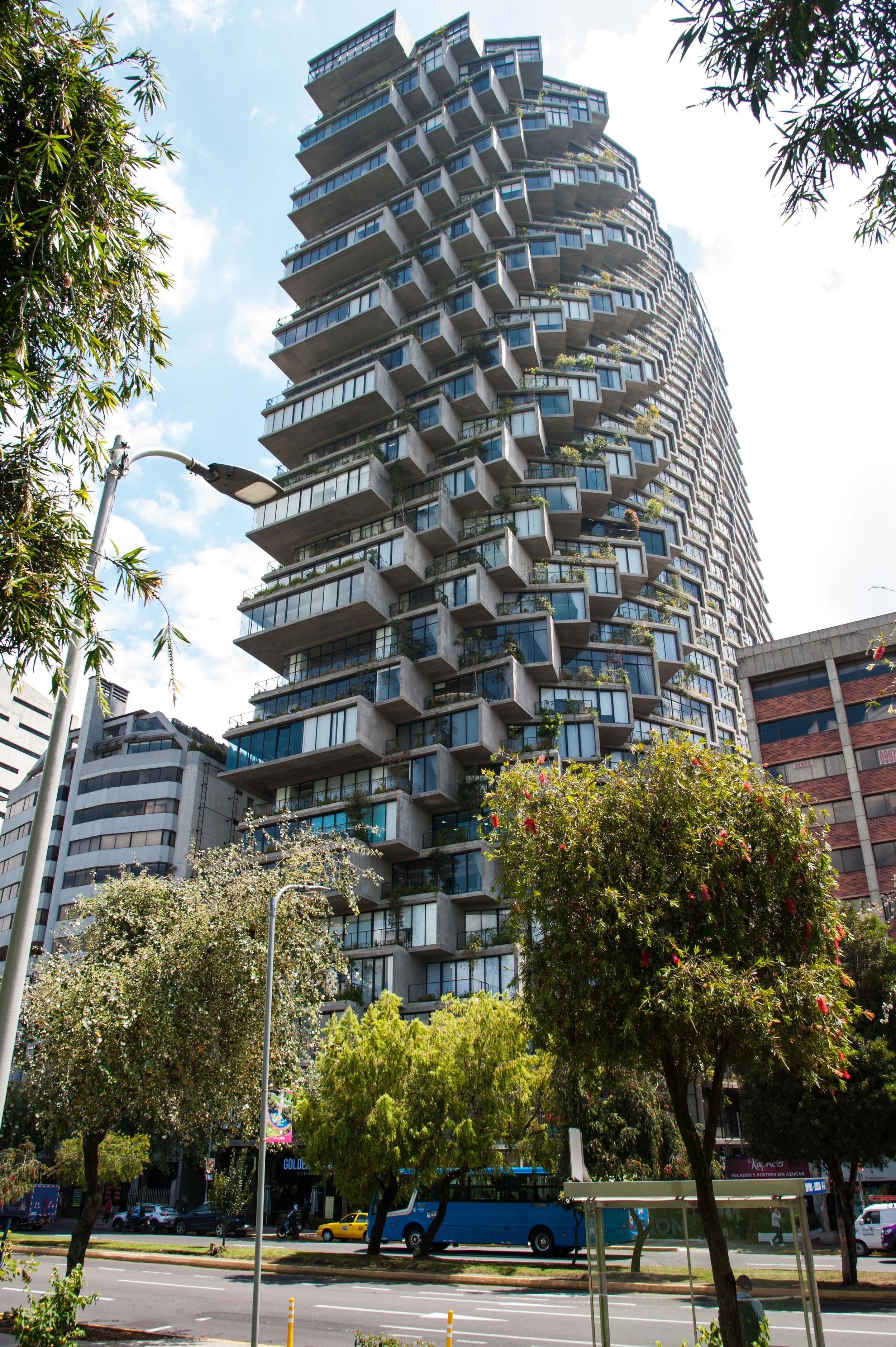 Tall modern residential building with protruding balconies and glass windows, surrounded by trees, on a sunny day with blue sky and clouds.