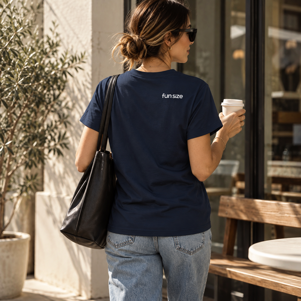 Woman with a black bag and sunglasses holding a coffee cup outside a café, wearing a navy blue t-shirt with 'fun size' text on the back and light blue jeans, with a plant and outdoor furniture nearby.