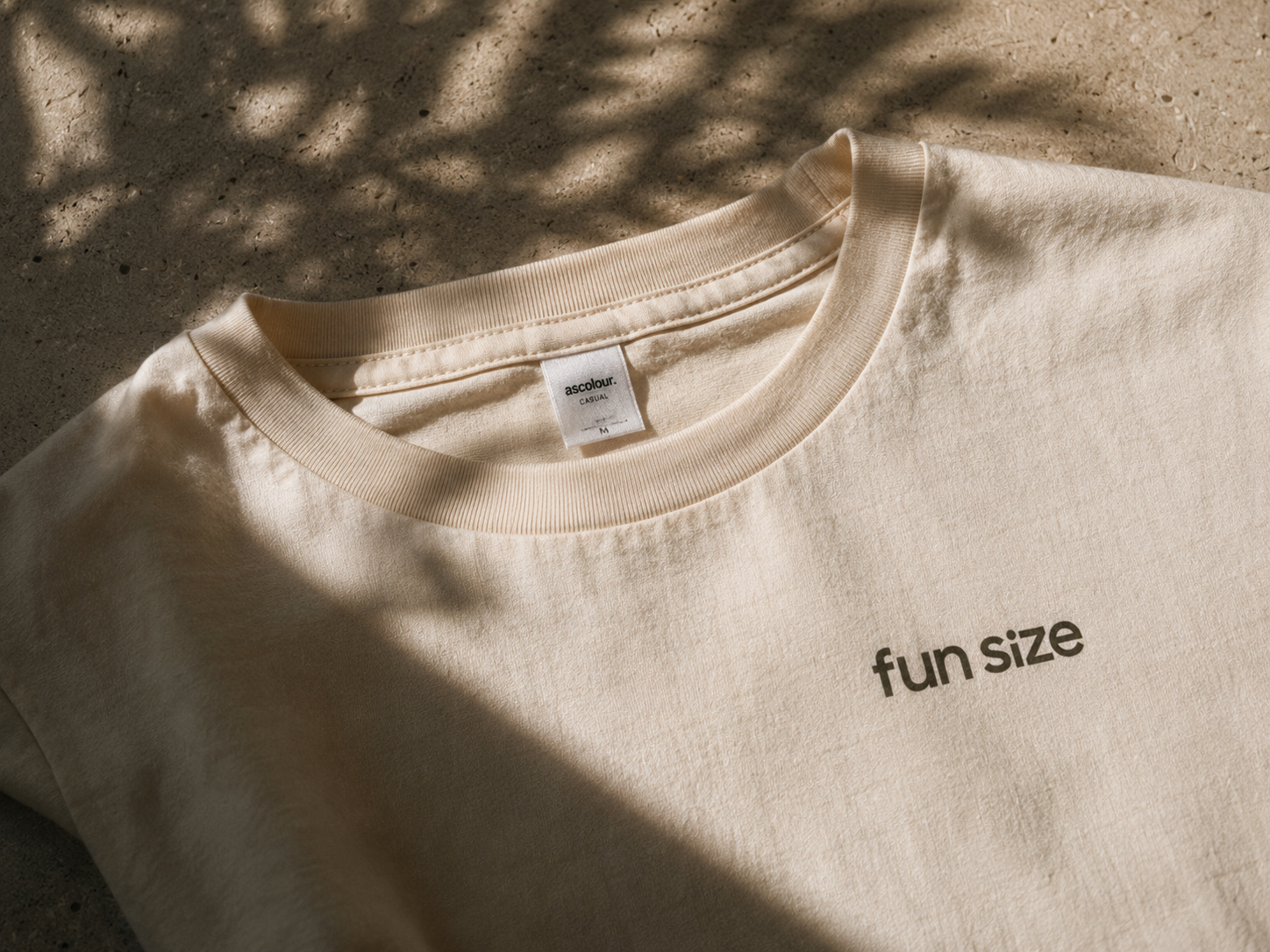 A cream-colored casual T-shirt with the words "fun size" printed in black on the front, laid flat on a textured surface with shadows cast over it.