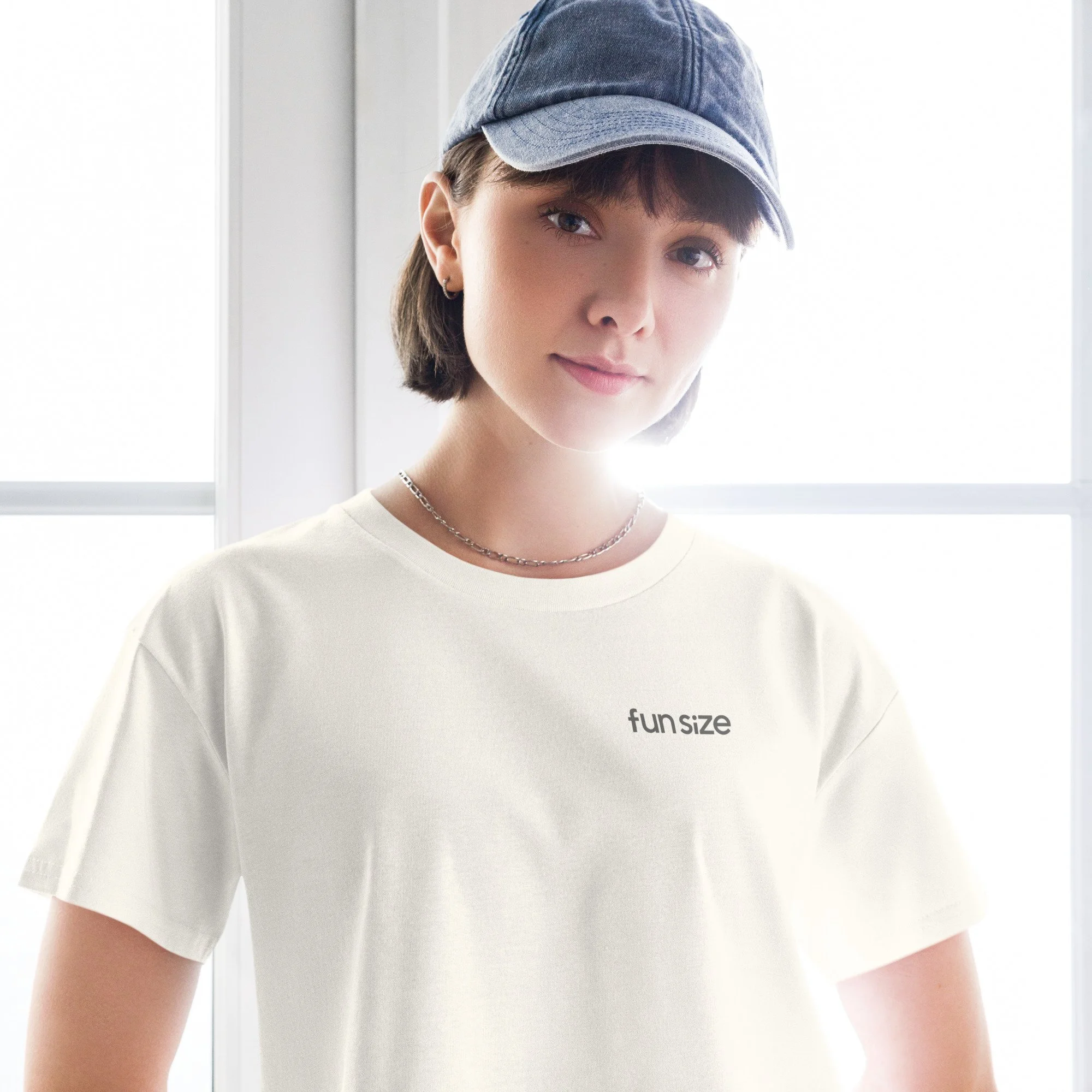 Close-up of a young woman wearing a light-colored FunSize t-shirt, silver chain necklace, hoop earrings, and a faded blue baseball cap, standing near a window with natural light.