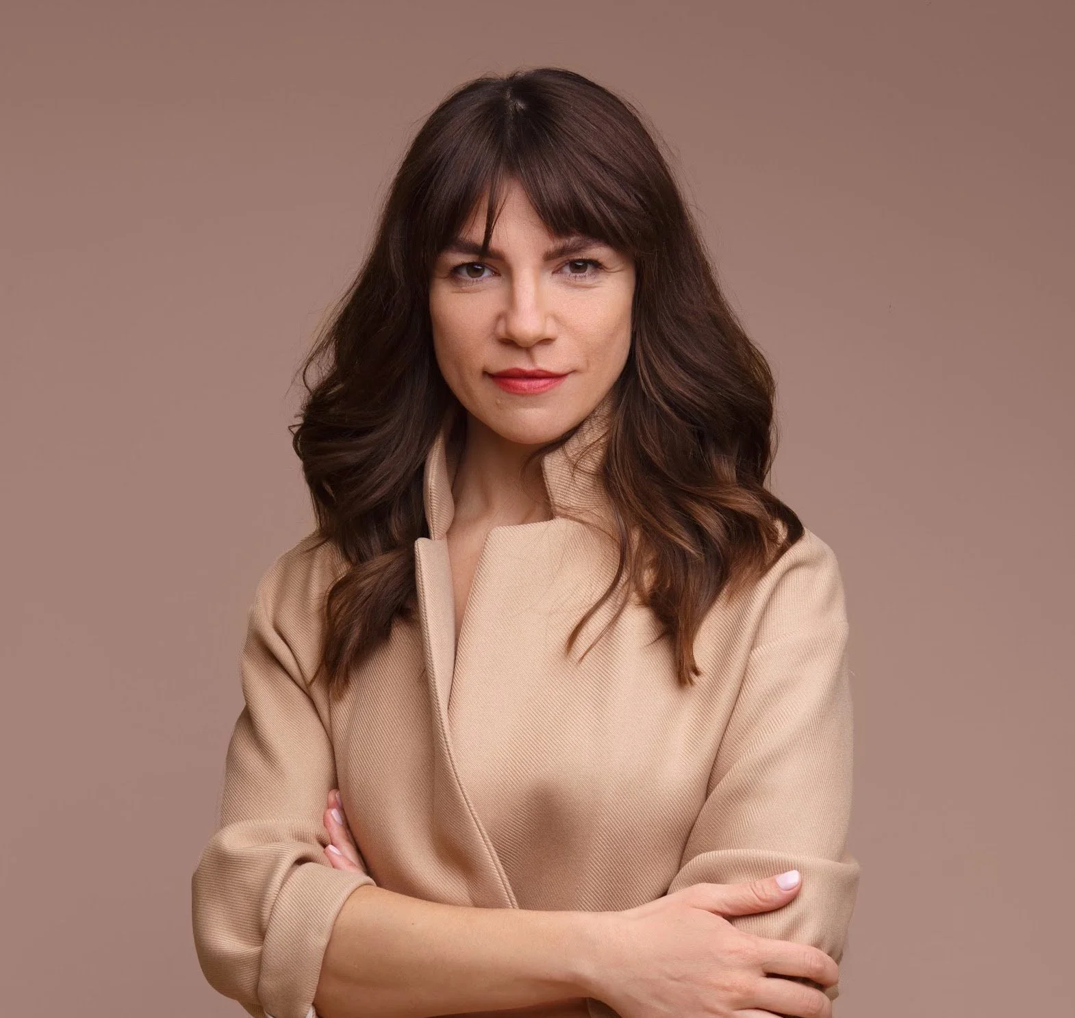 A woman with long brown hair styled in soft waves, wearing a beige top, stands against a plain, muted brown background, with her arms crossed and a slight smile.