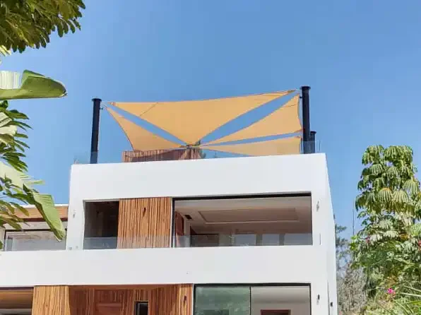 Vista superior de una casa moderna con una vela de sombra en la azotea, enmarcado por árboles verdes y un cielo azul claro.