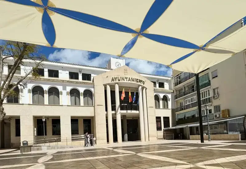 Edificio del ayuntamiento con banderas y velas de sombra cuadradas en la plaza central.