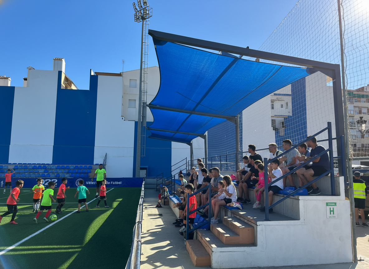Público en una grada de un campo de fútbol viendo un partido de niños en el campo, cubiertos y protegidos del sol por una vela de sombra azul