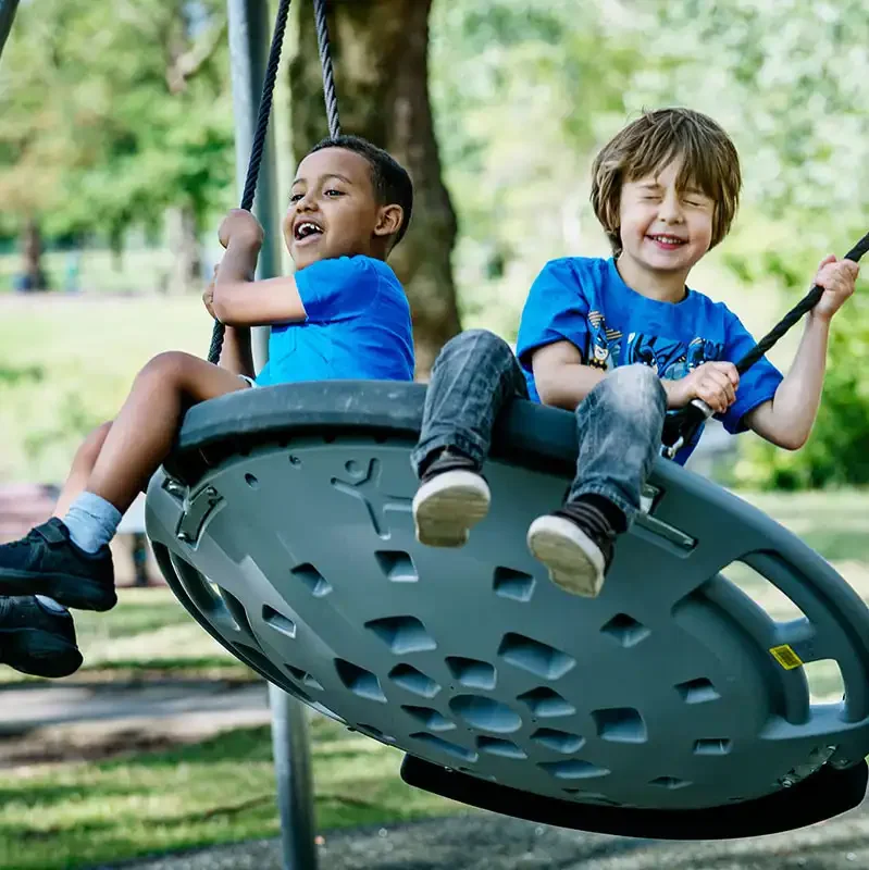 niños jugando en una hamaca circular con base de plastico, en un parque verde en una mañana de verano