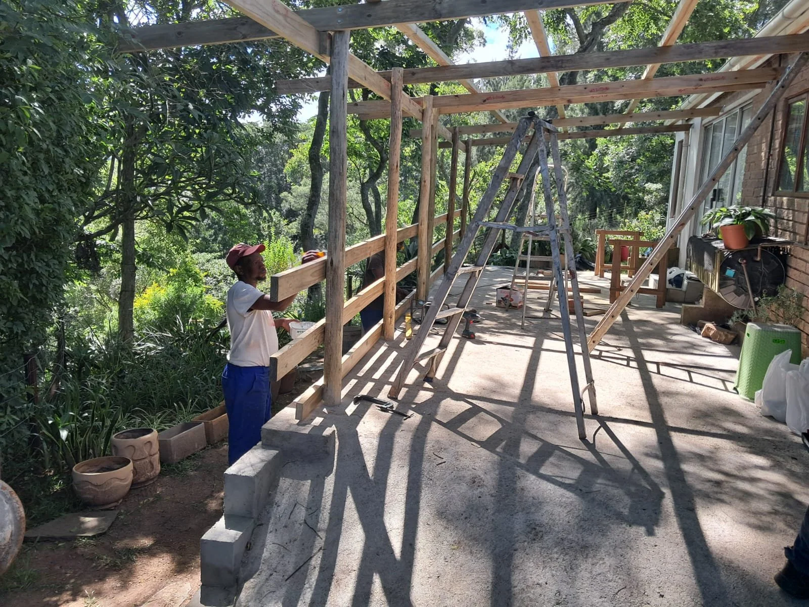 A person is working on building a wooden structure on a balcony or terrace, with trees and greenery in the background. Construction tools and materials.