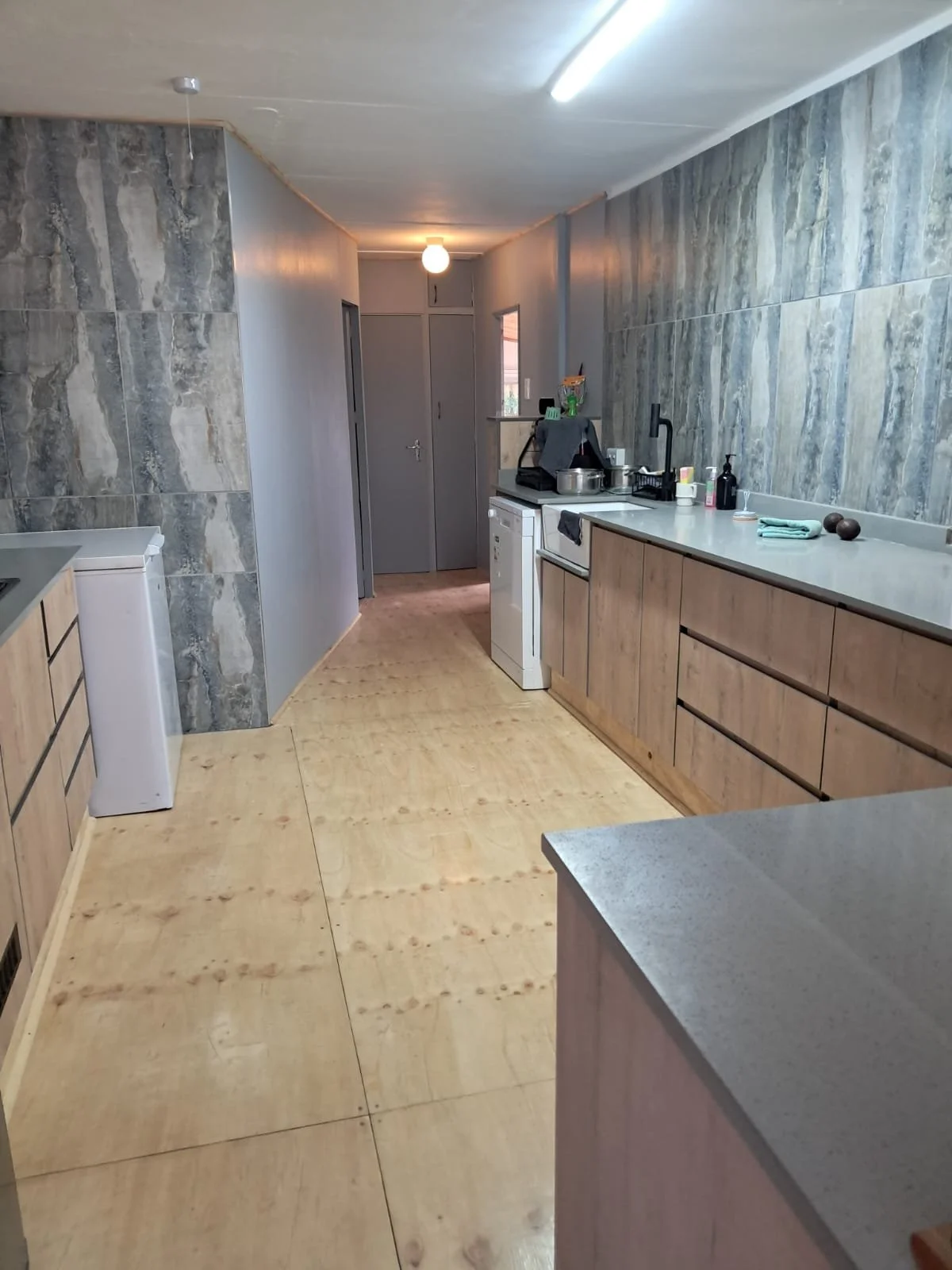 A kitchen with wood cabinets, a gray countertop, and a wall with gray marble tiles. The floor is made of treated plywood.
