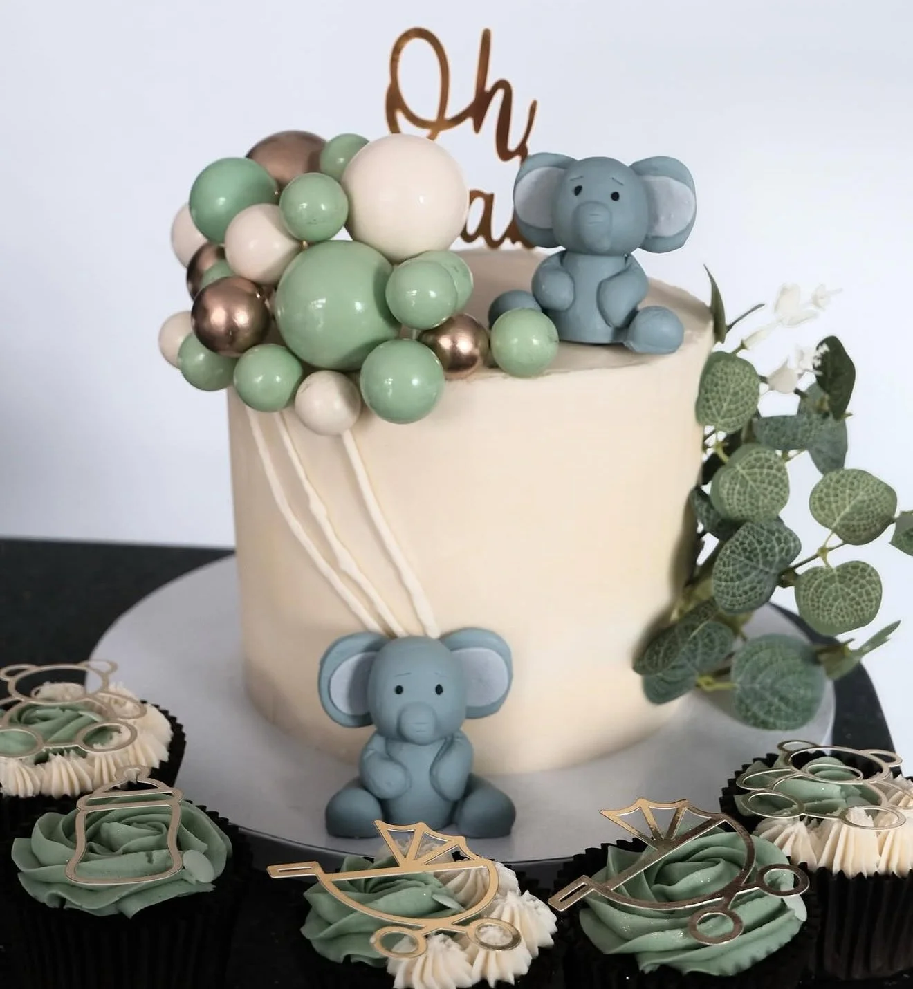 A cake decorated with blue elephant figurines, green and metallic-colored balloon decorations, green leaves, and a gold birthday topper with the word 'Oh' on it.
