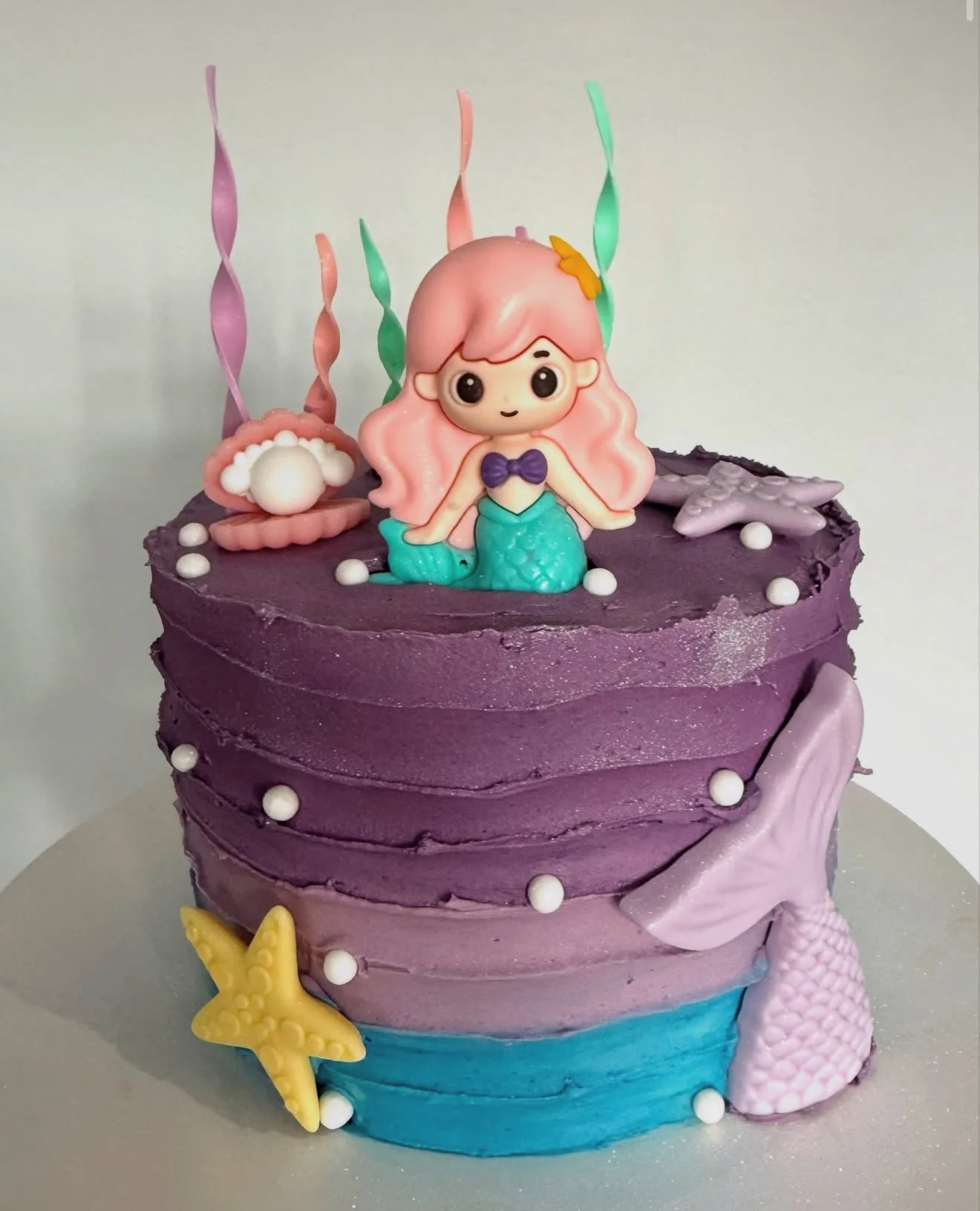 A two-layer celebration cake with purple and blue ocean-themed decorations, including a mermaid figurine with pink hair and a purple bikini top, shell-shaped decorations, starfish, and coral, topped with pink, green, and purple spiraled candles.