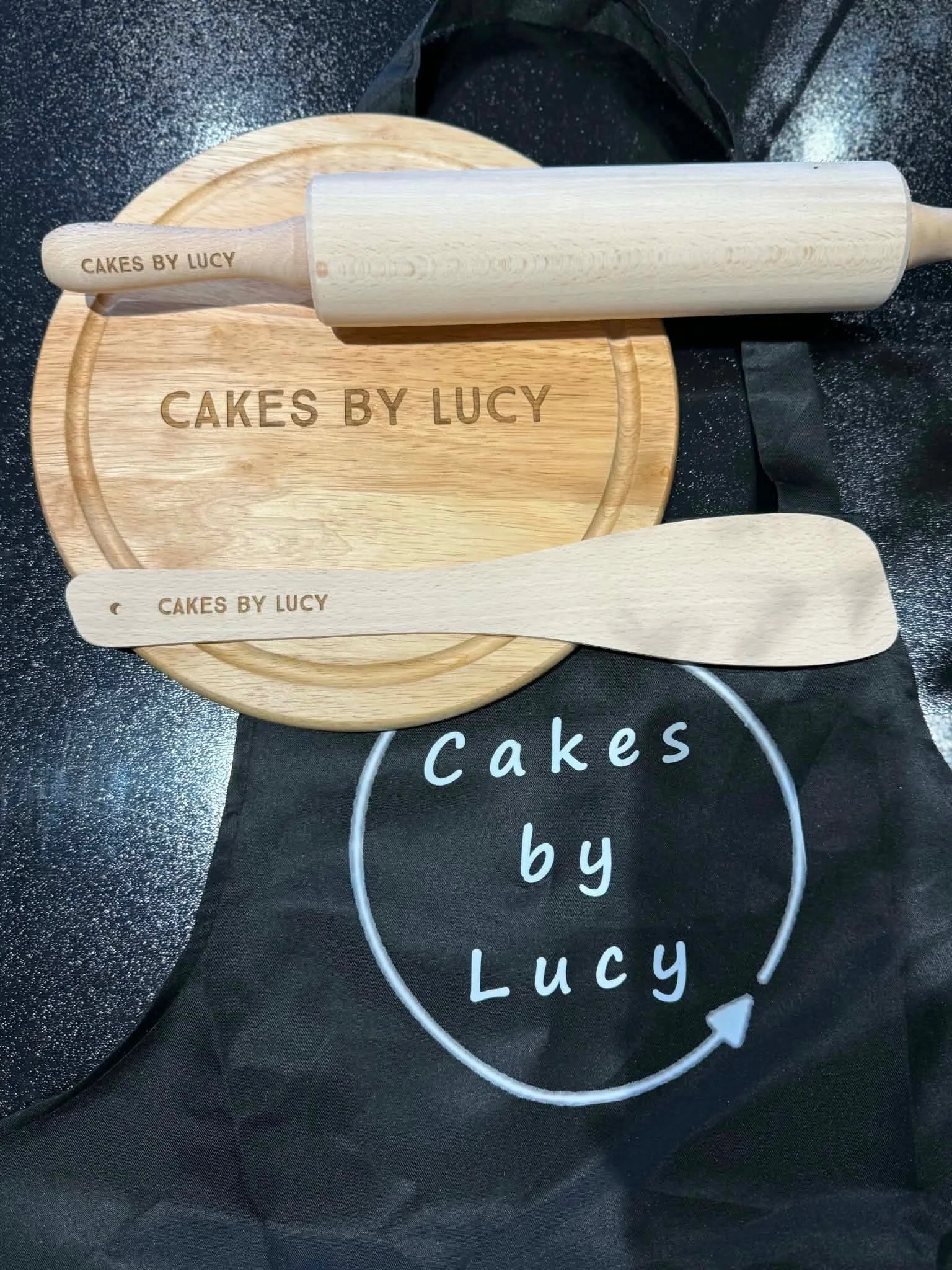 A wooden baking rolling pin engraved with 'CAKES BY LUCY' rests on a round wooden cutting board with the same inscription. The background includes a black surface with a white and green circular design that says 'Cakes by Lucy.'