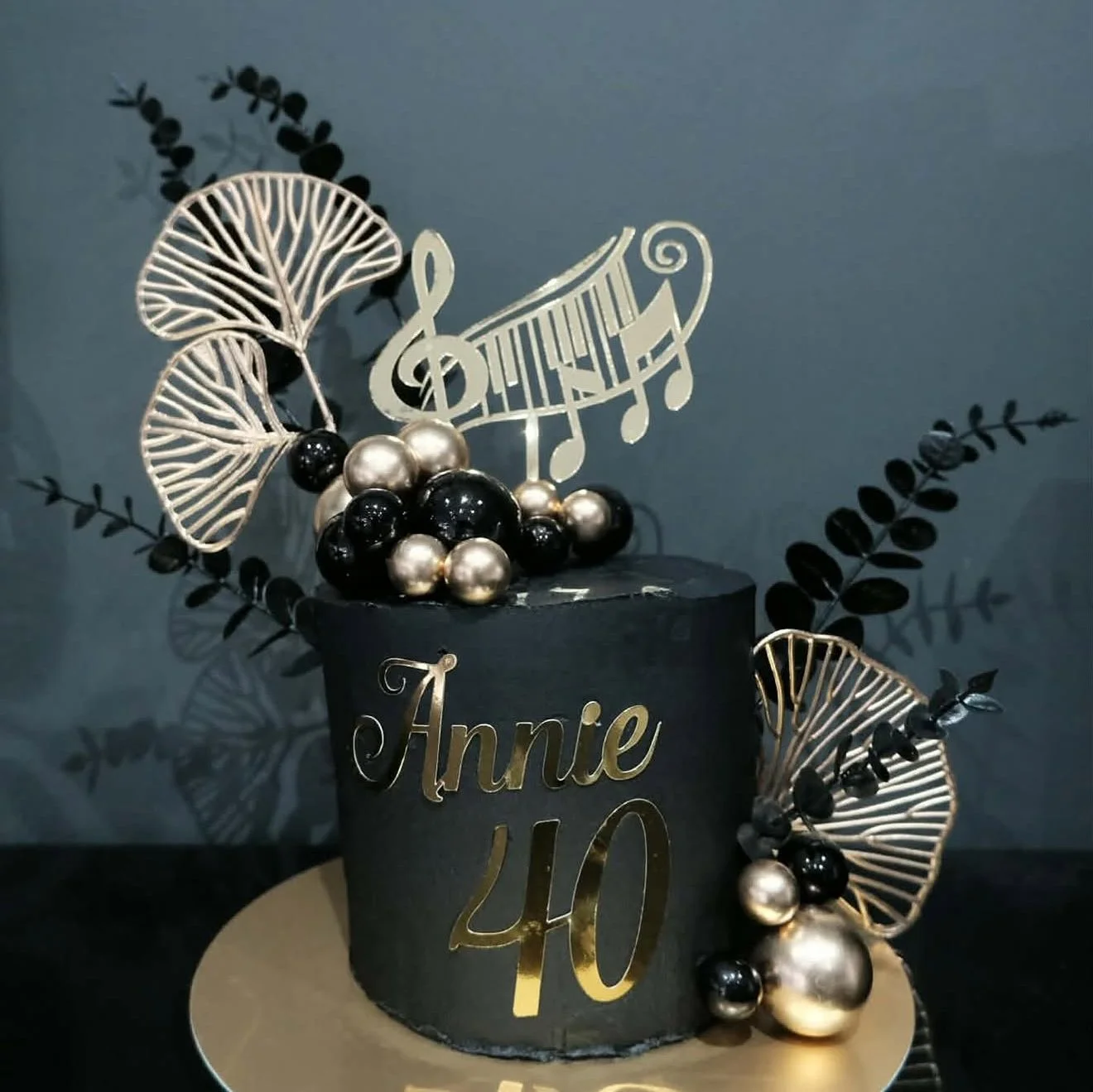 A black birthday cake decorated with black and silver balls, gold and silver leaves, a gold treble clef musical note, and a gold topper with the name 'Annie' and the number '40' in gold.