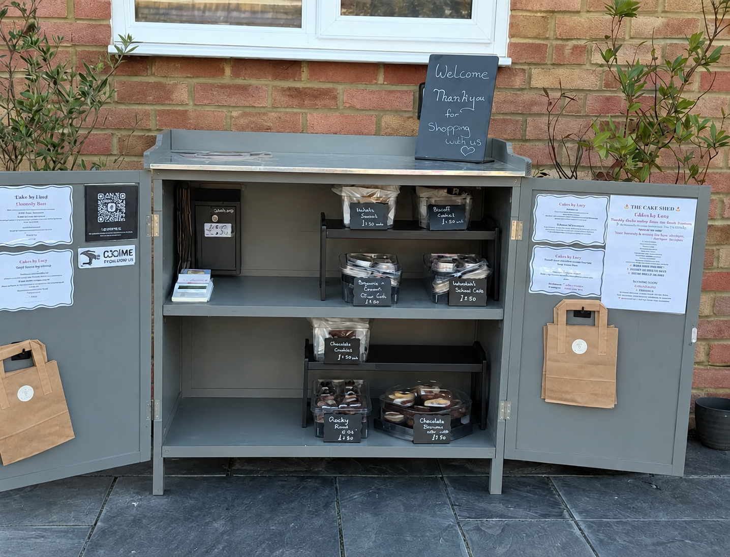 Outdoor display cabinet with baked goods for sale, including cookies, brownies, and cakes, with handwritten chalkboard signs and printed menus on the cabinet doors, against a brick wall background with potted plants.