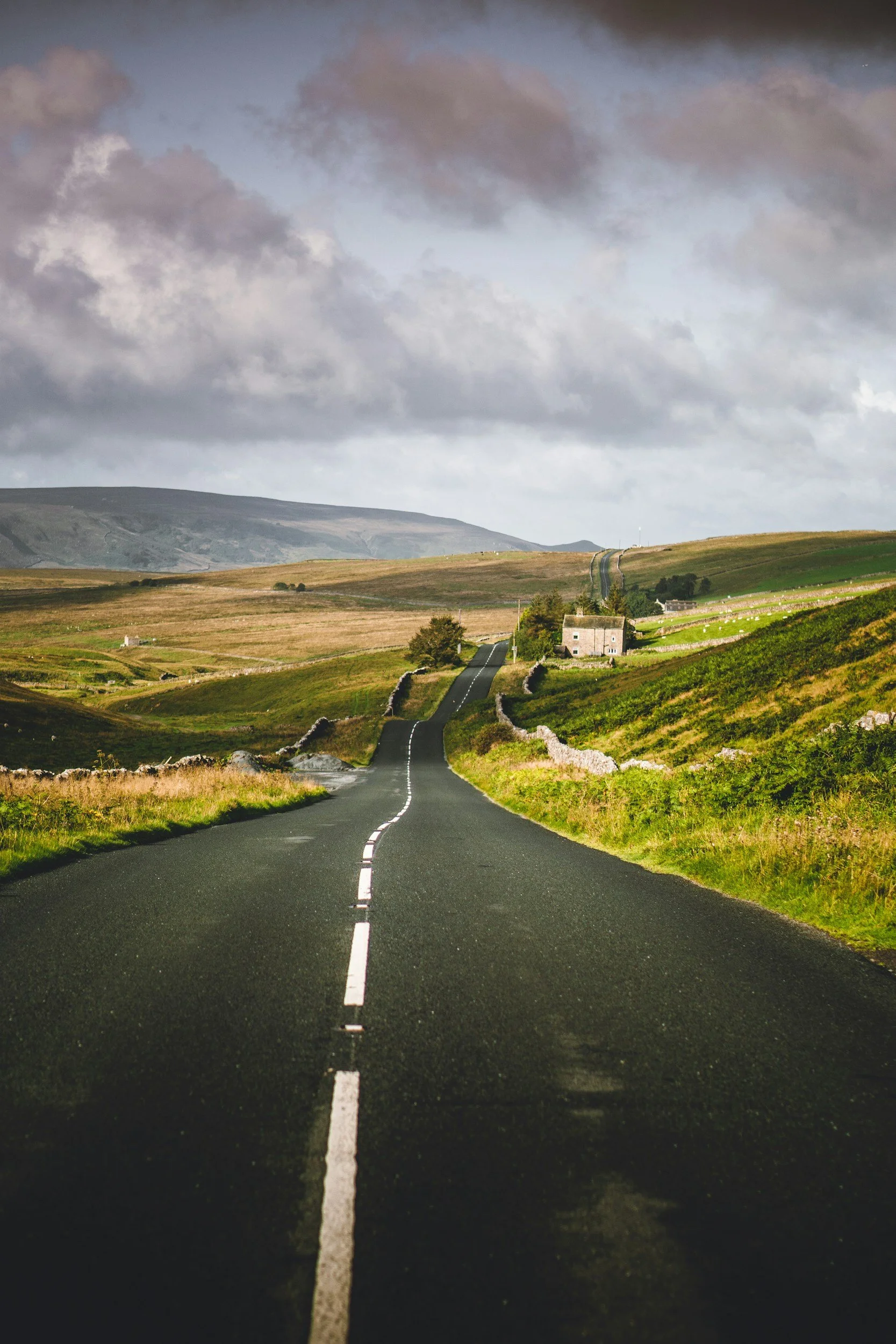 A long scenic country road running through green fields and hills under a cloudy sky.